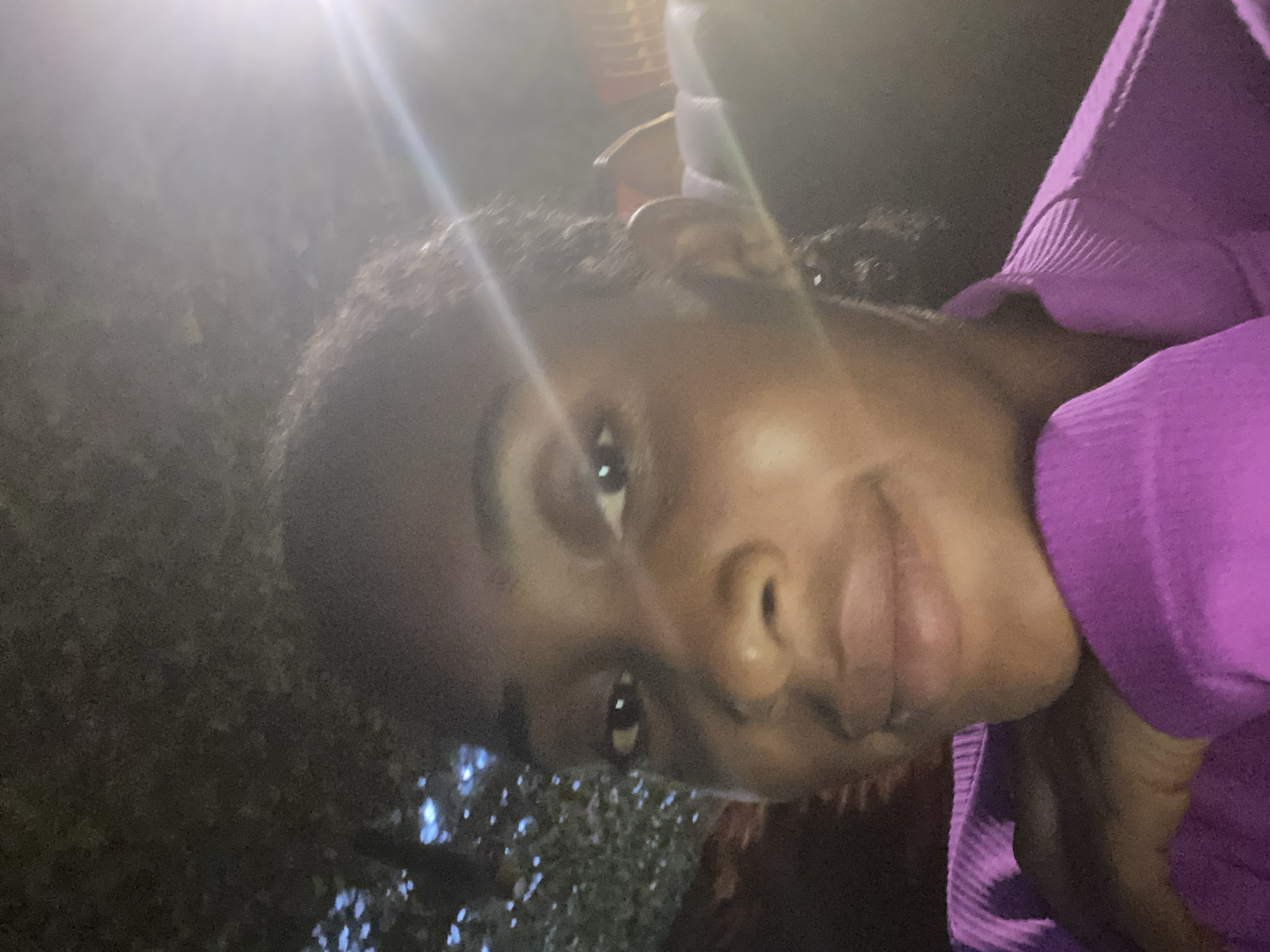 A young Black woman is pictured in a close-up, selfie-style shot, possibly taken outdoors at night. She has dark hair and is wearing a vibrant purple top with a ribbed texture. Her left hand is resting gently on her chin, and she is looking directly at the camera with a relaxed expression. In the background, faint lights and the silhouette of trees suggest an outdoor setting. A bright light source, possibly a street lamp or camera flash, is visible to the right, creating a glare across the image. The overall ambiance is informal and personal, with no specific indicators of a location within Abuja, Nigeria.