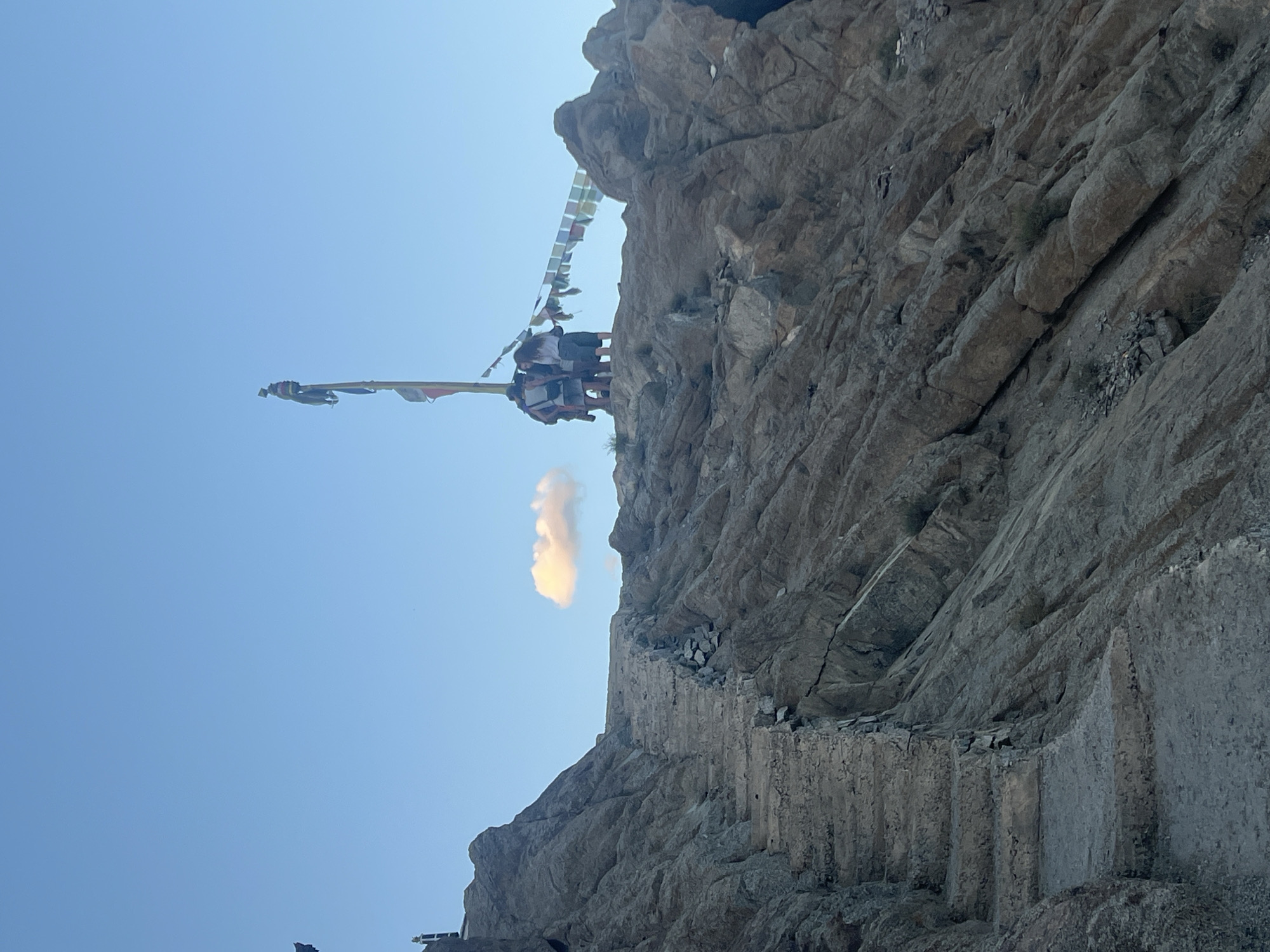 An outdoor, eye-level, full shot captures a mountainous scene under a clear blue sky with a single, small cloud. In the foreground, a stone staircase is carved into the side of a rocky incline, appearing weathered and somewhat broken in sections. The staircase leads upwards towards the summit of the mountain. At the top of the incline, a group of approximately five individuals are gathered around a tall, wooden pole adorned with prayer flags. The prayer flags are a mix of colors and are strung out horizontally to the right of the pole. The terrain is rugged and rocky, with evidence of erosion and fractured rock formations. The lighting suggests it is either early morning or late afternoon. The location is the Shanti Stupa in Leh, India.