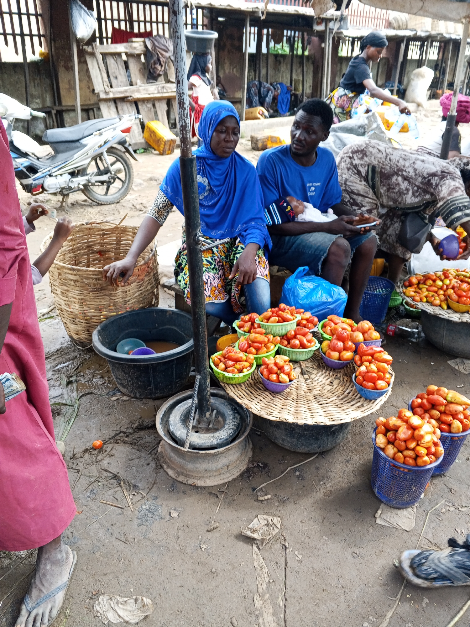 The image depicts a bustling market scene in Lokoja, Nigeria, with a focus on vendors and their produce. In the foreground, a woman in a blue hijab and patterned skirt is tending to a display of tomatoes. Beside her, a man in a blue t-shirt holds a sleeping infant. The ground is a mix of dirt and mud, with various baskets and containers holding tomatoes. A motorcycle is parked in the background, suggesting a typical mode of transport in the area. Other market-goers and stalls are visible, contributing to the lively atmosphere. The scene appears to be an outdoor market, characteristic of many urban centers in Nigeria.