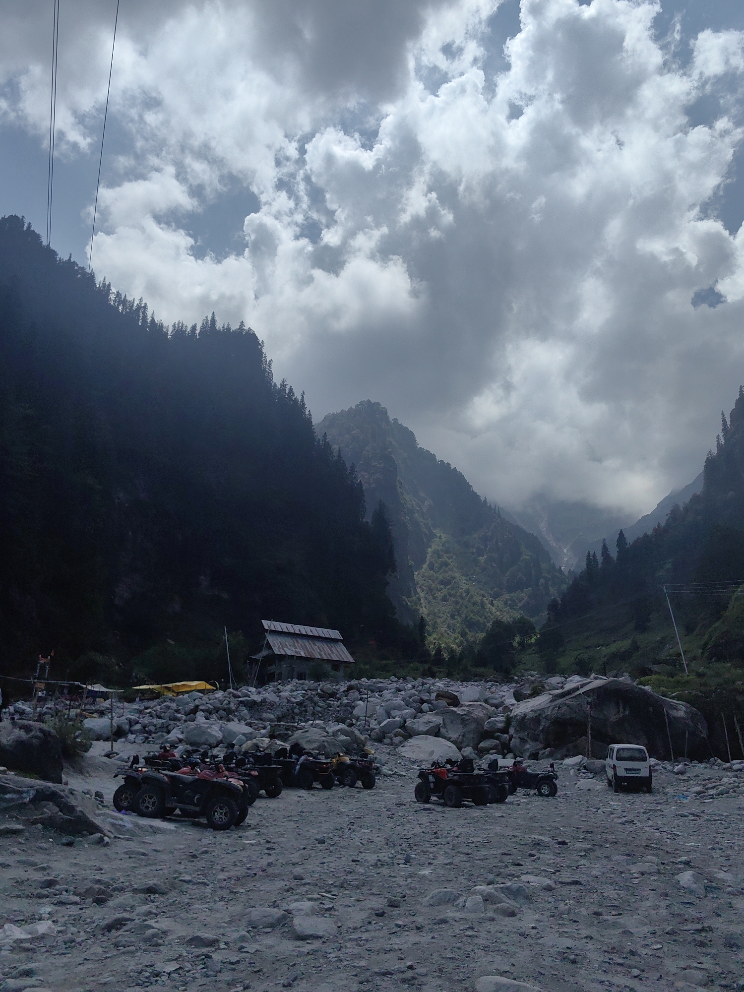 The image shows a wide, rocky riverbed in a mountainous valley, likely in the vicinity of Majhach, India, based on the context. Several ATVs are parked on the gravelly ground in the foreground. In the middle ground, a rustic, dilapidated building with a corrugated metal roof stands near the base of a steep, tree-covered mountainside. The background features a dramatic mountain landscape with a narrow valley opening up, partially obscured by clouds and mist. The sky is filled with a mixture of dark, heavy clouds and brighter patches, suggesting dynamic weather conditions. The overall scene depicts a rugged, natural environment with signs of human activity in the form of parked vehicles and a small structure.