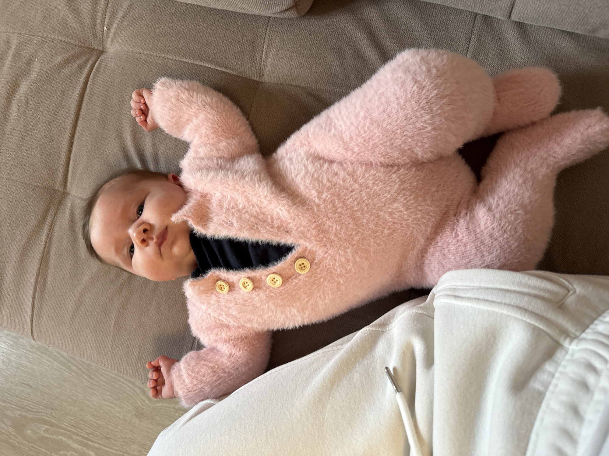 A baby lies on a tan sofa. The baby is wearing a pink, fuzzy, button-down onesie over a black shirt. A person wearing white sweatpants sits next to the baby. The location is presumed to be in the city of Chaikovsky, Russia.