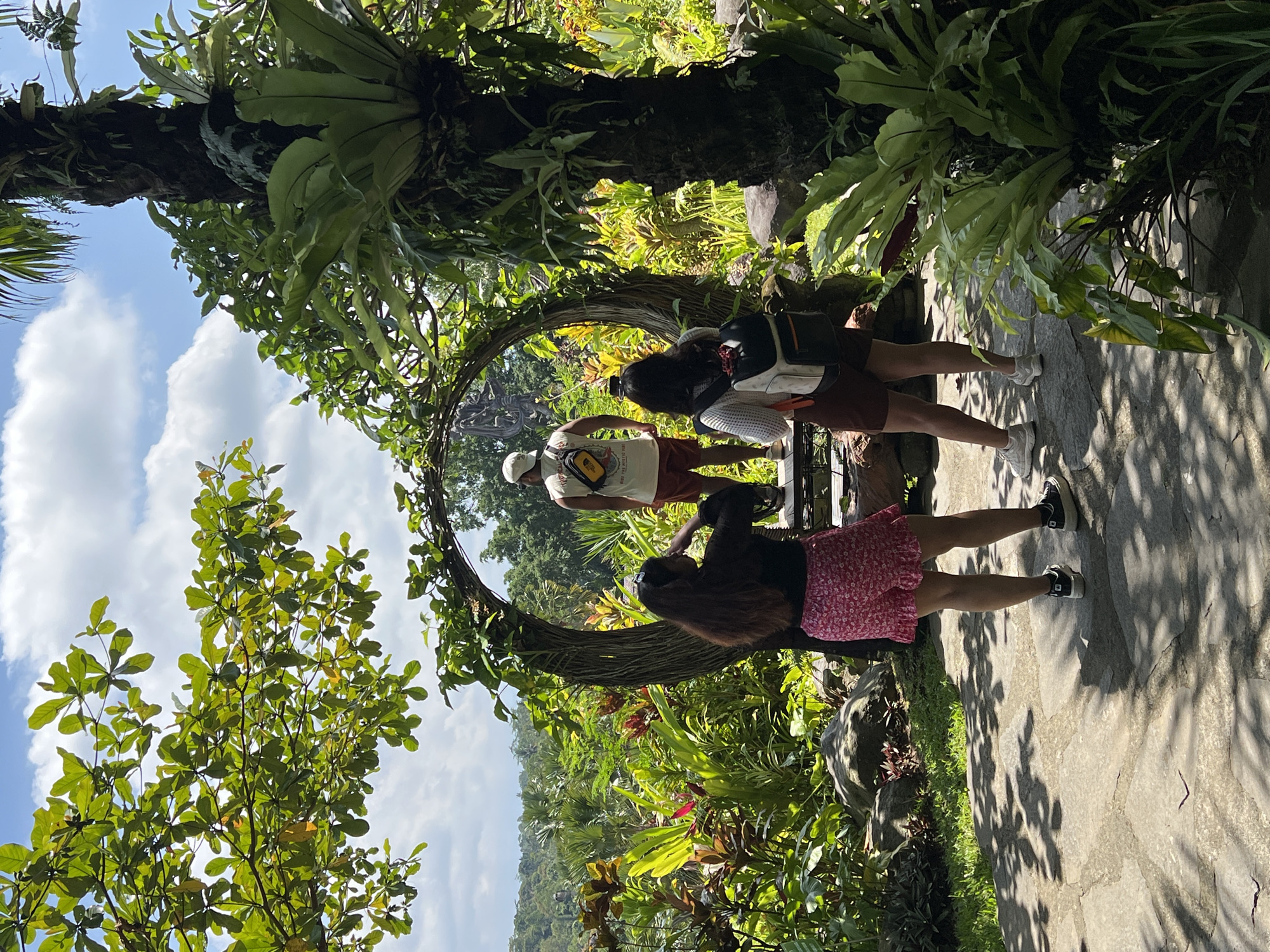 Three individuals stand within a circular, vine-covered structure positioned on a stone path amidst lush vegetation, under a partly cloudy sky. The location is Kedewatan, Indonesia. One individual, wearing a white shirt and cap, faces away from the camera. Another individual wears a dark top and a pink floral skirt. The third individual has a backpack. The surrounding environment is filled with diverse plants and trees.