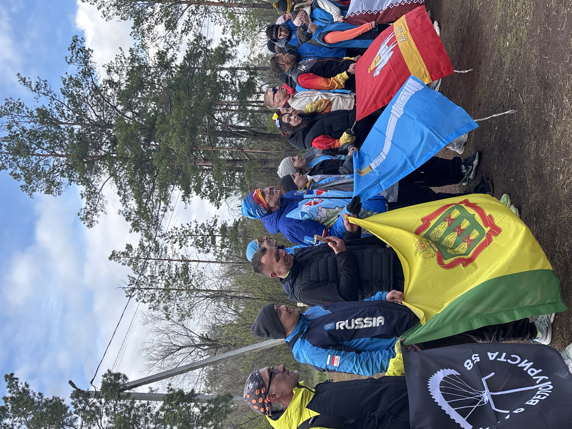 The image depicts a gathering of people outdoors, likely for a sporting or outdoor event. A group of adults, predominantly men, are visible standing or sitting, dressed in casual and sportswear. The background features tall pine trees and a dirt ground, suggesting a forest edge or park setting. The sky is partly cloudy with patches of blue.
Several flags are prominently displayed among the participants. From left to right (as seen from the front of the group):
1. A large yellow and green flag with a detailed coat of arms featuring three golden sheaves of wheat against a green field, framed by a red border, which is the flag of Penza Oblast, Russia.
2. A bright blue flag with a white, stylized pattern that resembles a mammoth tusk or traditional ornament, identifying it as the flag of the Yamal-Nenets Autonomous Okrug, Russia.
3. Partially visible behind the blue flag is a red and yellow flag with a white, stylized animal, which appears to be the flag of the Republic of Kalmykia, Russia.
One participant is wearing a blue and black jacket with "RUSSIA" clearly printed on the chest and sleeve. Another person wears a black hat with the text "G. TRAIL". At the bottom of the frame, a black flag is visible, featuring a white bicycle wheel graphic and the Cyrillic text "ВЕЛОТУРИСТА 58" (VELOTURISTA 58), which translates to "Bicycle Tourist 58". The number "58" corresponds to the vehicle registration code for Penza Oblast, suggesting this is a local cycling club or event. Power lines are faintly visible against the sky in the upper left. The natural lighting indicates it is daytime. The combination of regional flags and sportswear suggests a multi-regional sporting event, possibly a cycling race or tour, taking place in Russia.