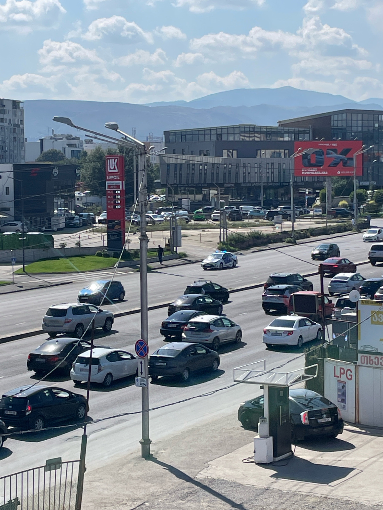 This is an aerial, eye-level, medium shot of a city street in Tbilisi, Georgia, on a sunny day. The foreground features a gas station with an LPG sign and a black Toyota Prius being refueled. The street is busy with traffic, including several cars, a police car with blue flashing lights, and a few pedestrians walking on the sidewalk. In the background, there are modern buildings, mountains, and a clear blue sky with scattered clouds. The image captures a typical urban scene with elements of daily life and infrastructure.