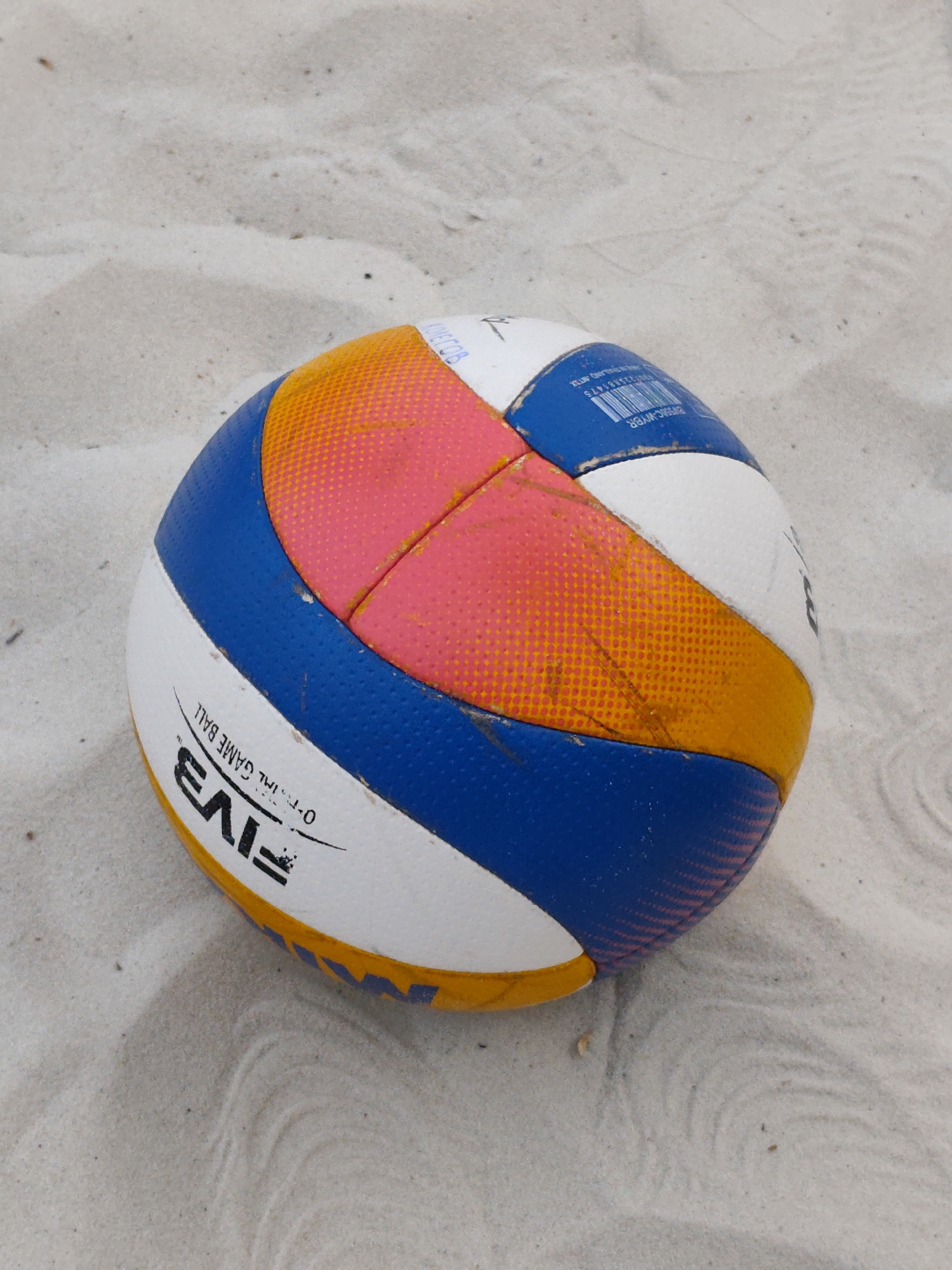 The image shows a FIVB-branded volleyball lying on light-colored sand. The ball is multi-colored, featuring white, blue, orange, and pink panels. The sand appears fine and uniform, suggesting a beach or sand pit setting. The image was not taken in the city of Chaikovsky, Russia. The city of Chaikovsky is a city in the Perm Krai region of Russia, but it is not on the beach.