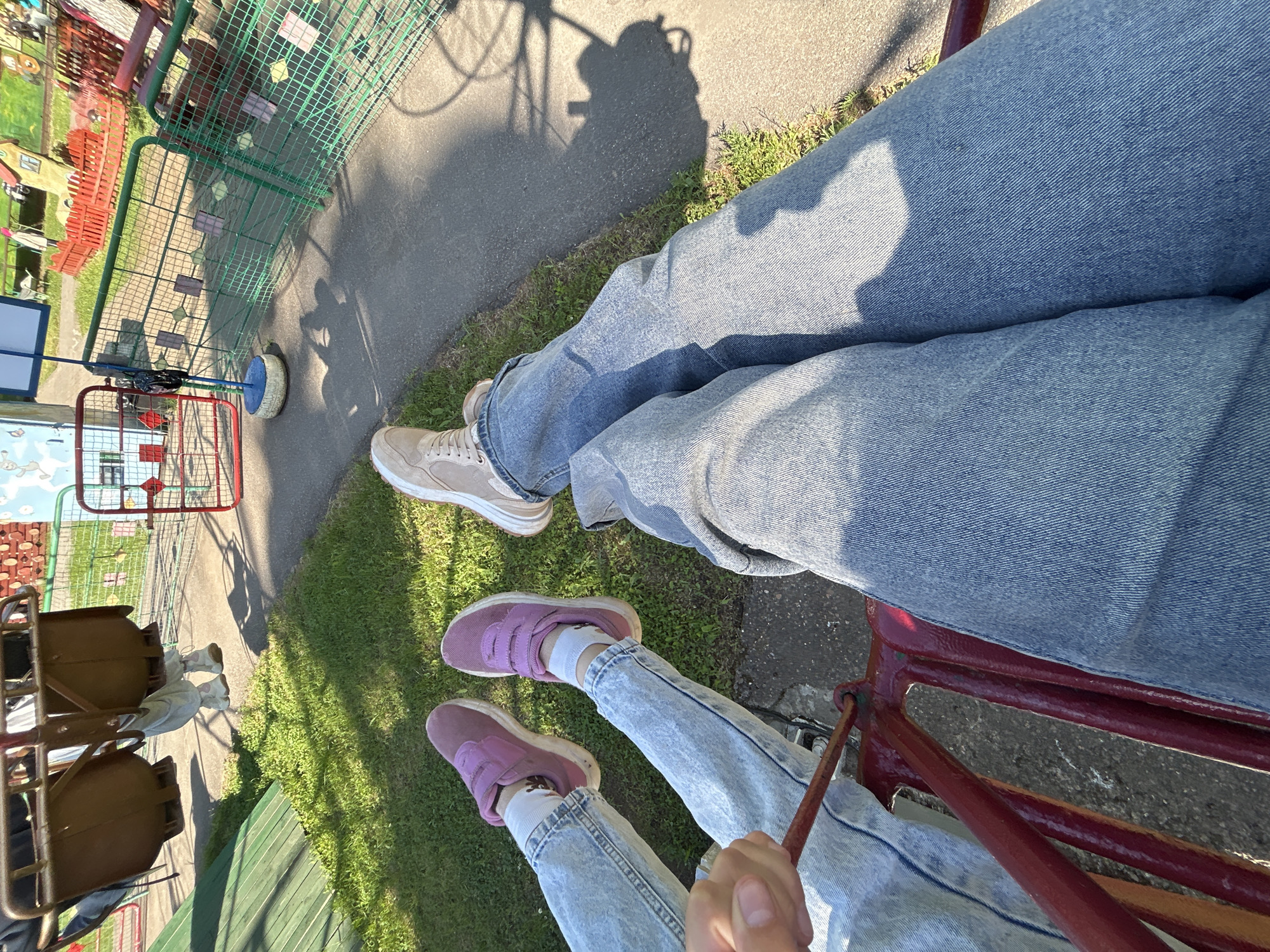 Two individuals are depicted from an overhead perspective, presumably seated on a ride in an outdoor setting. The individual in the foreground wears light blue jeans and pink sneakers with white socks. Their hand is visible, holding onto a metal bar. The other person wears darker blue jeans and beige sneakers. The ride appears to be a swing-type carousel. A green fence with red accents is visible in the background, along with a paved surface and grass. The shadow of the carousel's structure is cast on the paved ground. This appears to be taken in Chaikovsky, Russia, given the original request context.
