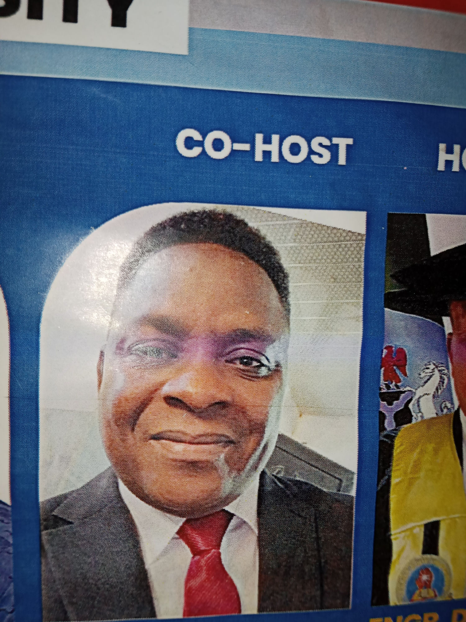 The image shows a close-up of two men in formal wear, seemingly at an event. The man on the left is in focus, wearing a dark suit, a white shirt, and a red tie. He has dark skin and is smiling. Above his portrait, the words "CO-HOST" are printed in white capital letters on a blue background. To his right, another man's face is partially visible, wearing a black cap and a yellow sash. Behind the man on the left is a textured white surface. The image appears to be a photograph of a poster or banner, possibly displayed at a university or conference event in Jalingo, Nigeria, given the context of the event name.