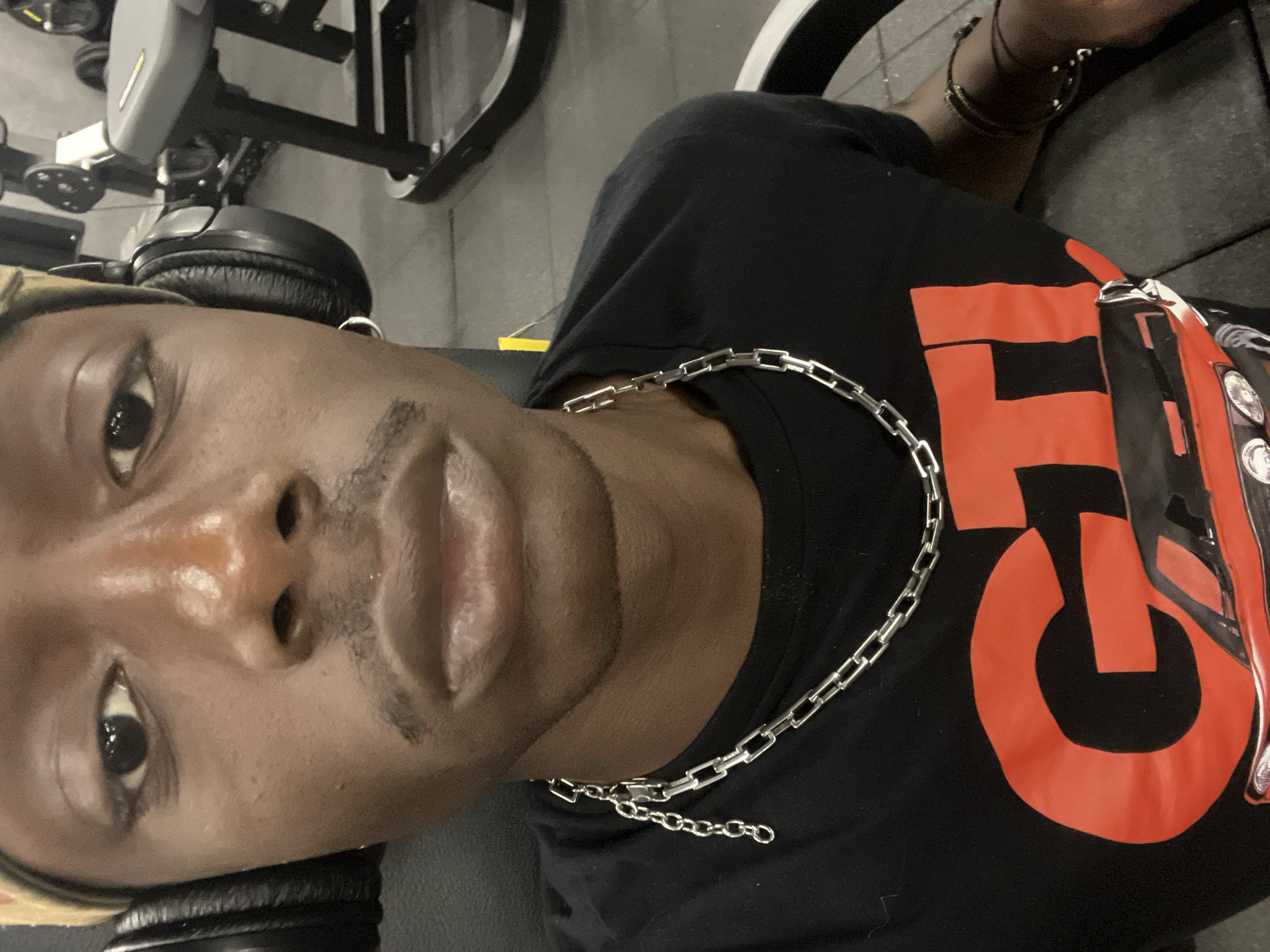 A selfie of a dark-skinned person wearing headphones, a silver chain necklace, and a black t-shirt with a red design. The person is indoors, likely in a gym setting, as exercise equipment is visible in the background. The location is not discernible as Barcelona, Spain, based on the image.
