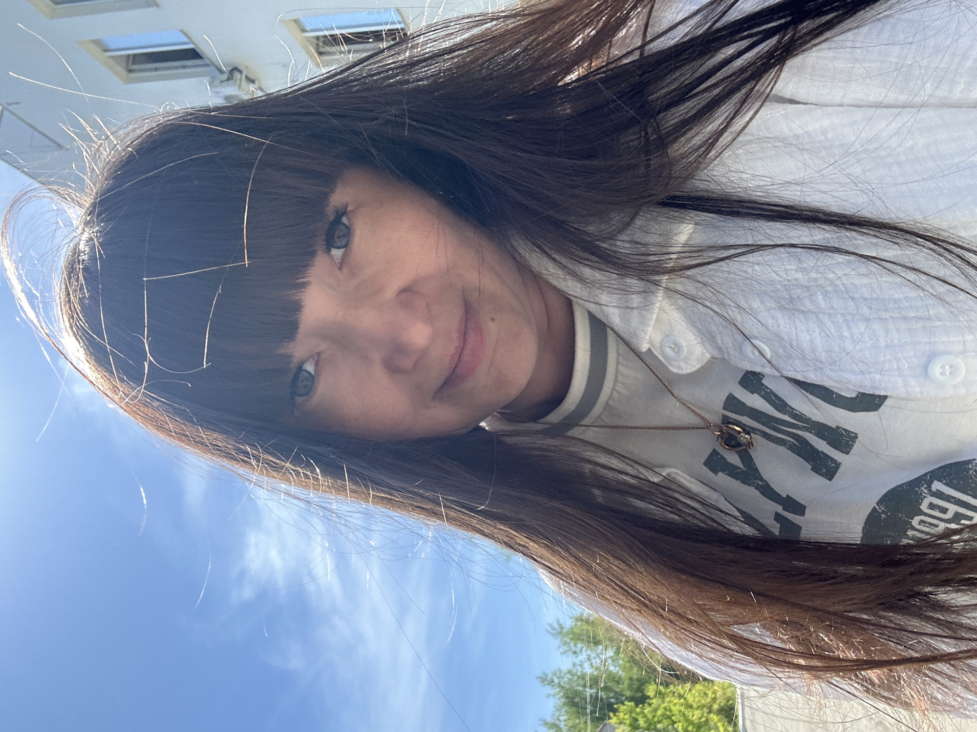 A woman with brown hair and bangs is looking at the camera from a low angle. She is wearing a white button-down shirt over a striped-collar shirt and a necklace. The background includes a blue sky with some clouds, a building with windows, and green trees. The location is inferred to be Chaikovsky, Russia, based on the prompt instructions, but this is not evident from the visual information.