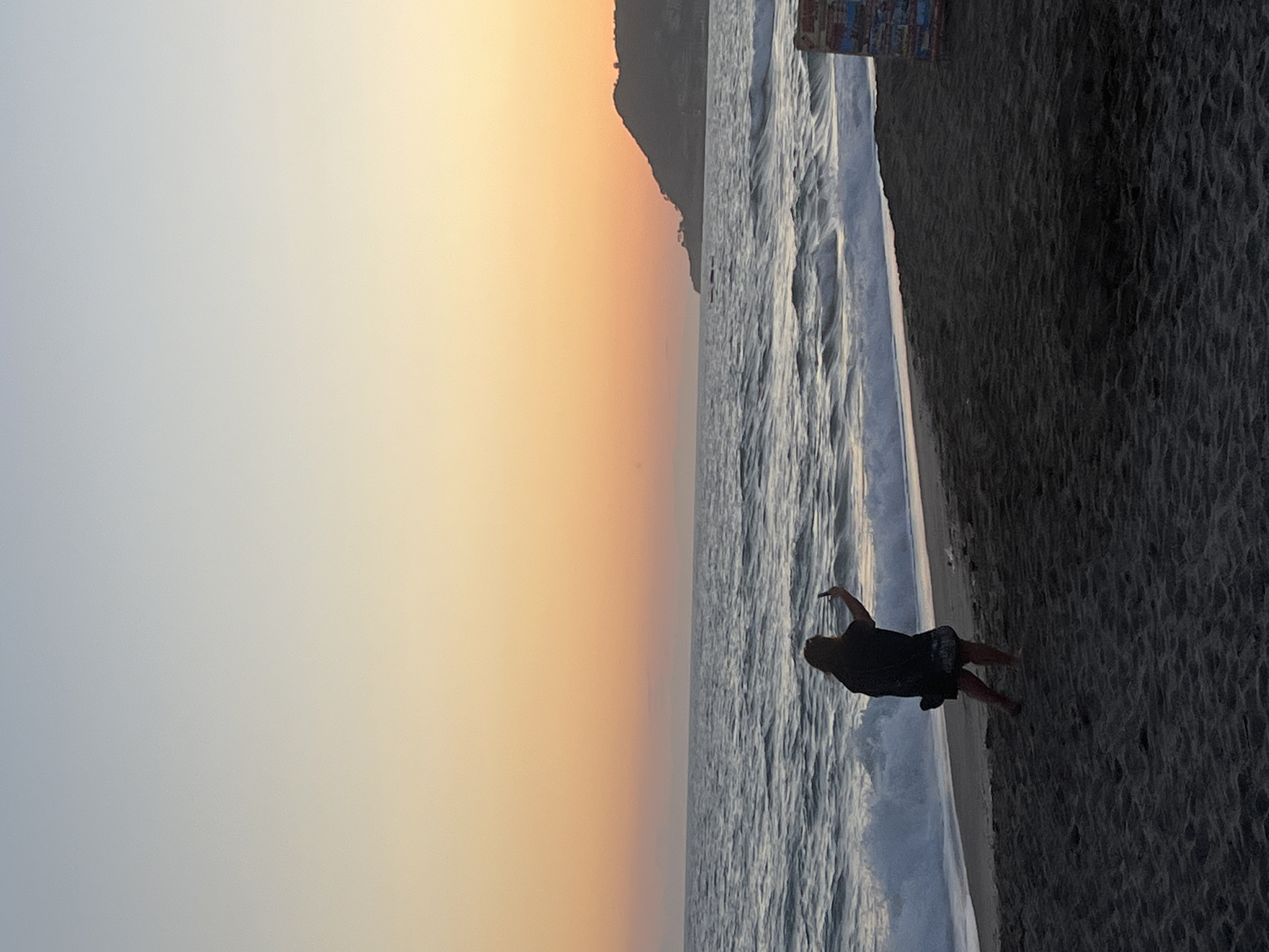 A person is standing on a sandy beach as waves crash in the sea at Alanya, Turkey. The person is wearing dark clothing. A land mass is visible along the horizon. The sky has gradations of color, from light blue to orange, suggesting dawn or dusk. The beach is dark and appears somewhat coarse.