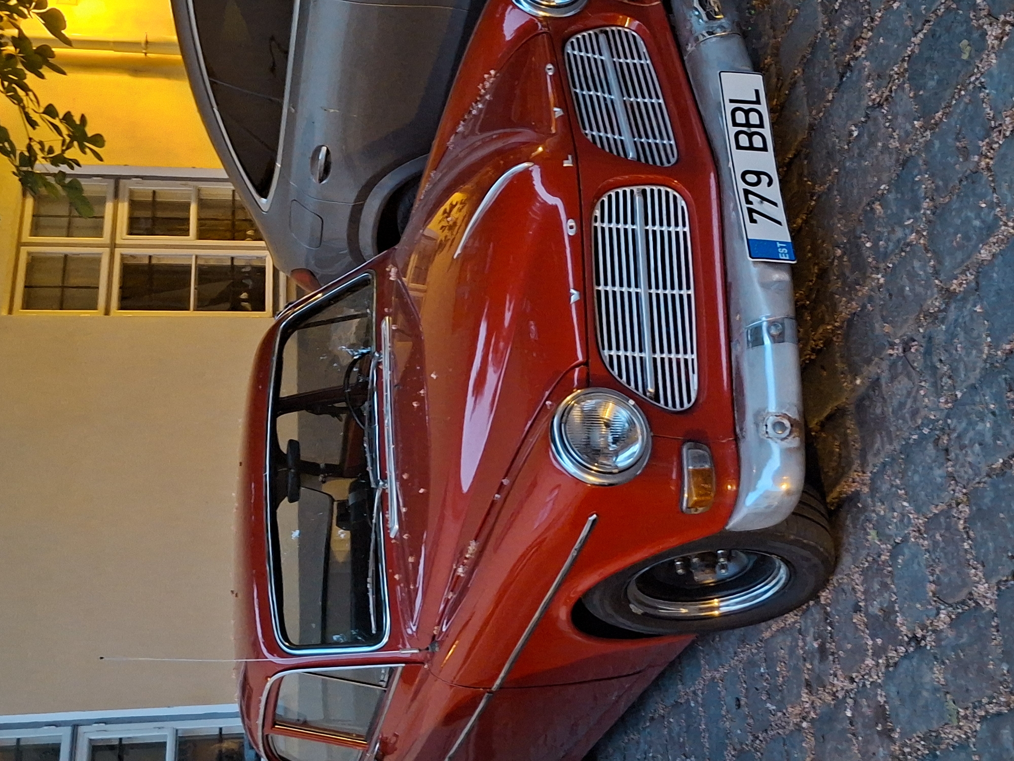 A red Volvo sedan, likely a P1800 model from the 1960s, is parked on a cobblestone street in Tallinn, Estonia. The car has a chrome grille and bumper, and a license plate that reads "779 BBL" with "EST" indicating its Estonian registration. The car's hood is slightly dirty with what appear to be small leaves or debris. In the background, a modern grey SUV is parked behind the Volvo. A building with large windows, possibly a historic structure, is visible to the left and right of the vehicles, with its facade illuminated by warm, yellow light, suggesting it is either evening or a streetlamp is on. The street is paved with dark, textured cobblestones. The scene appears to be a quiet street in an urban setting.