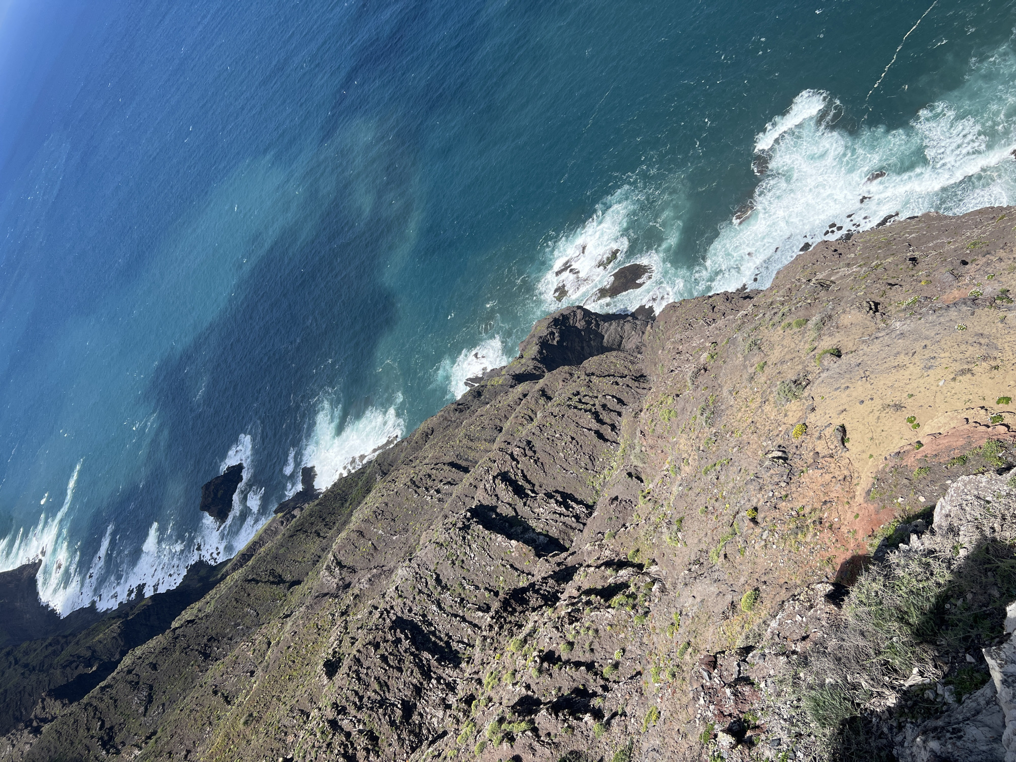 The image shows an aerial view of the coastline near Tirma, Spain. The Atlantic Ocean stretches into the distance, with waves crashing against the rocky shore. A steep, terraced cliff face dominates the lower portion of the image, with visible vegetation clinging to the rocks. The overall scene depicts a rugged, natural landscape where the land meets the sea.