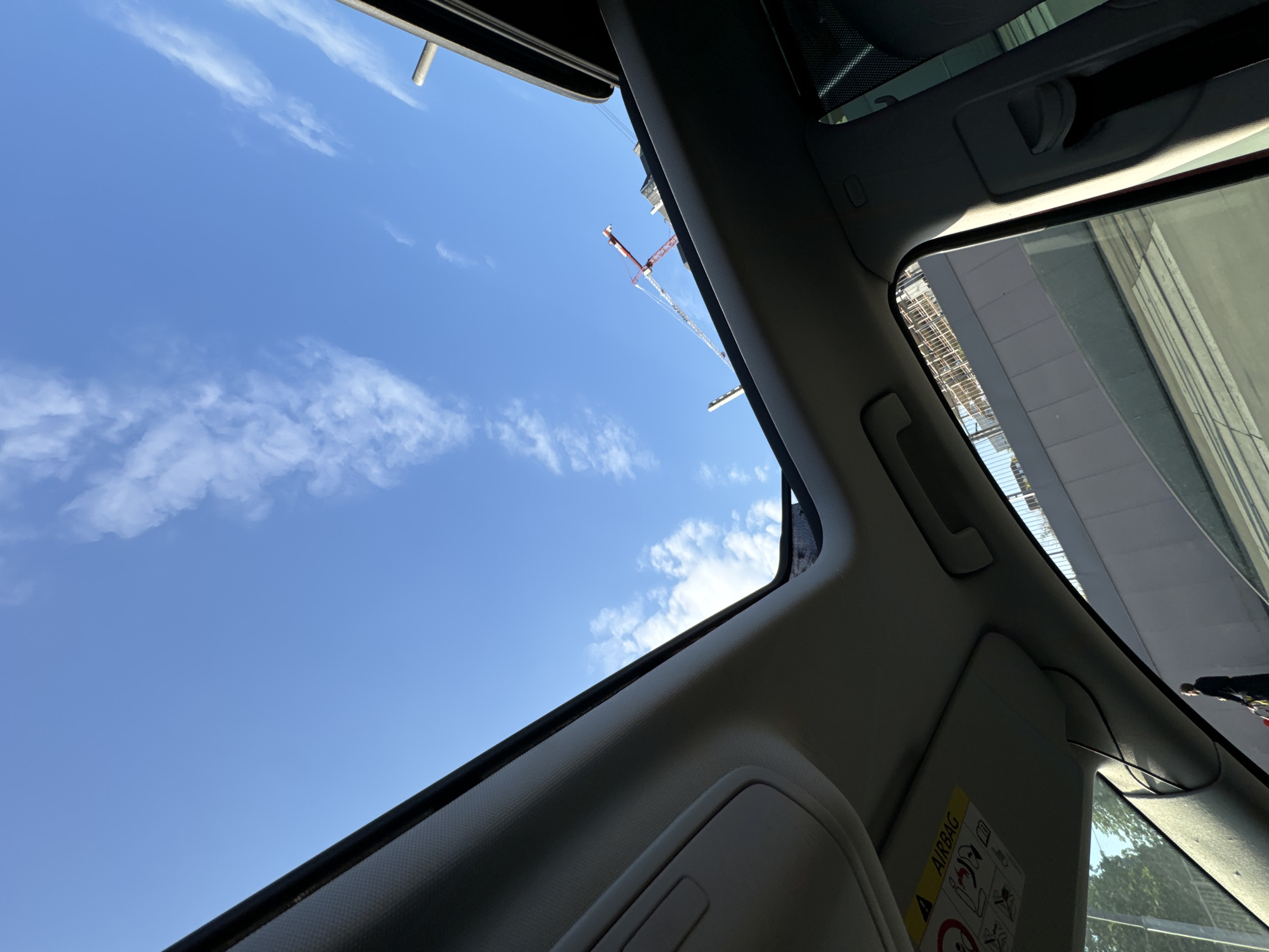 The image shows a view through an open car sunroof, looking upwards at a bright blue sky with some wispy clouds. A construction crane is visible in the distance. Part of a building with a scaffolding-like structure is also seen. The car's interior, including the sun visor and parts of the roof lining, frames the view. The picture appears to be taken during daylight hours. The presence of construction cranes and a building under construction could be in Moscow, Russia.