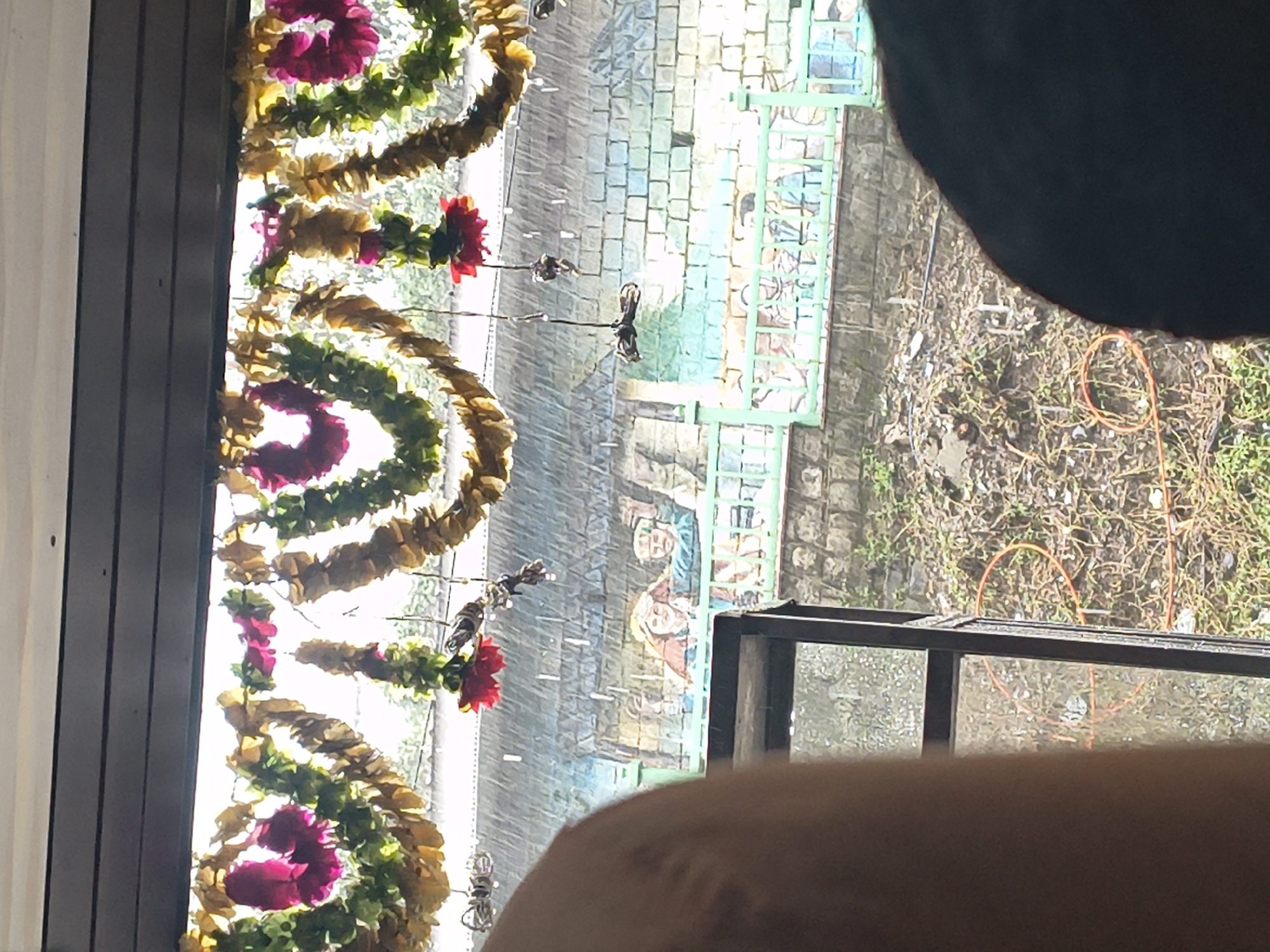 The image shows a view from indoors looking out through a window onto a rainy scene in Dharamshala, India. In the foreground, a portion of a person's shoulder and head, or possibly the interior of a vehicle, is visible on the left and right sides. Above the window, decorative garlands made of green leaves and dried flowers are hung. Outside the window, it is raining, with visible streaks of rain and water droplets.
In the middle ground, a mural or painting is visible on a brick wall, depicting several people. A green railing is in front of the mural. The area behind the railing appears to be a slope covered in vegetation and some orange circular objects, possibly hoses or ropes. The overall environment suggests a slightly overcast and wet day in a mountainous region, consistent with Dharamshala's climate.