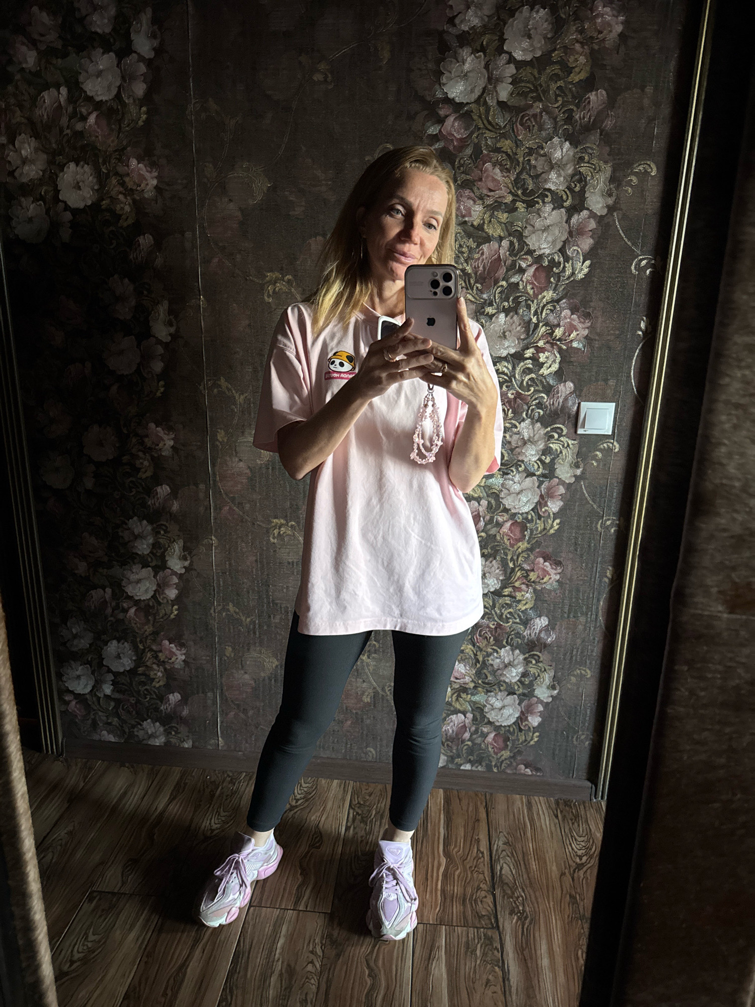 A blonde woman is taking a mirror selfie in a room. She is wearing a light pink t-shirt with a panda graphic on the left chest, black leggings, and light purple athletic shoes. She is holding a smartphone in her right hand and has a pink beaded charm attached to it. The background features floral wallpaper and a wooden floor. The scene appears to be indoors, possibly in a home or hotel room. No specific location in Torzhok, Russia can be determined from the image.