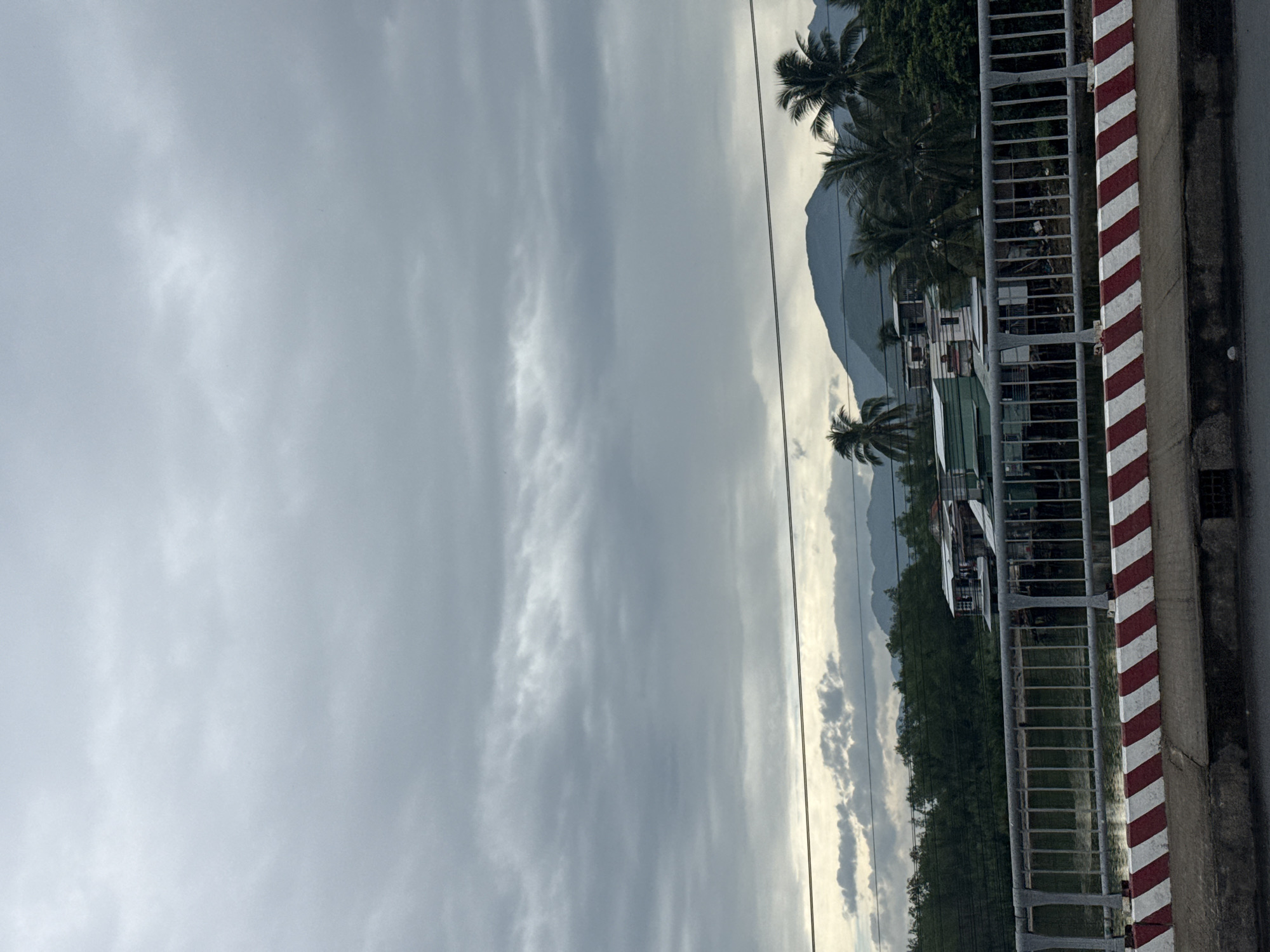 The image depicts a view of a canal or river in Nha Trang, Vietnam, likely from a bridge. The sky is heavily overcast with grey clouds, suggesting an impending or ongoing rain. In the background, lush green trees line the banks, interspersed with a cluster of buildings, some appearing to be houses on stilts over the water. Palm trees are visible amongst the foliage, indicating a tropical climate. Further in the distance, rolling hills or mountains are shrouded in the hazy atmosphere. In the foreground, the edge of the bridge is visible with a metal railing and a distinct red and white striped barrier. The overall scene is subdued due to the cloudy weather, with a focus on the natural landscape and the modest human habitation along the waterway.