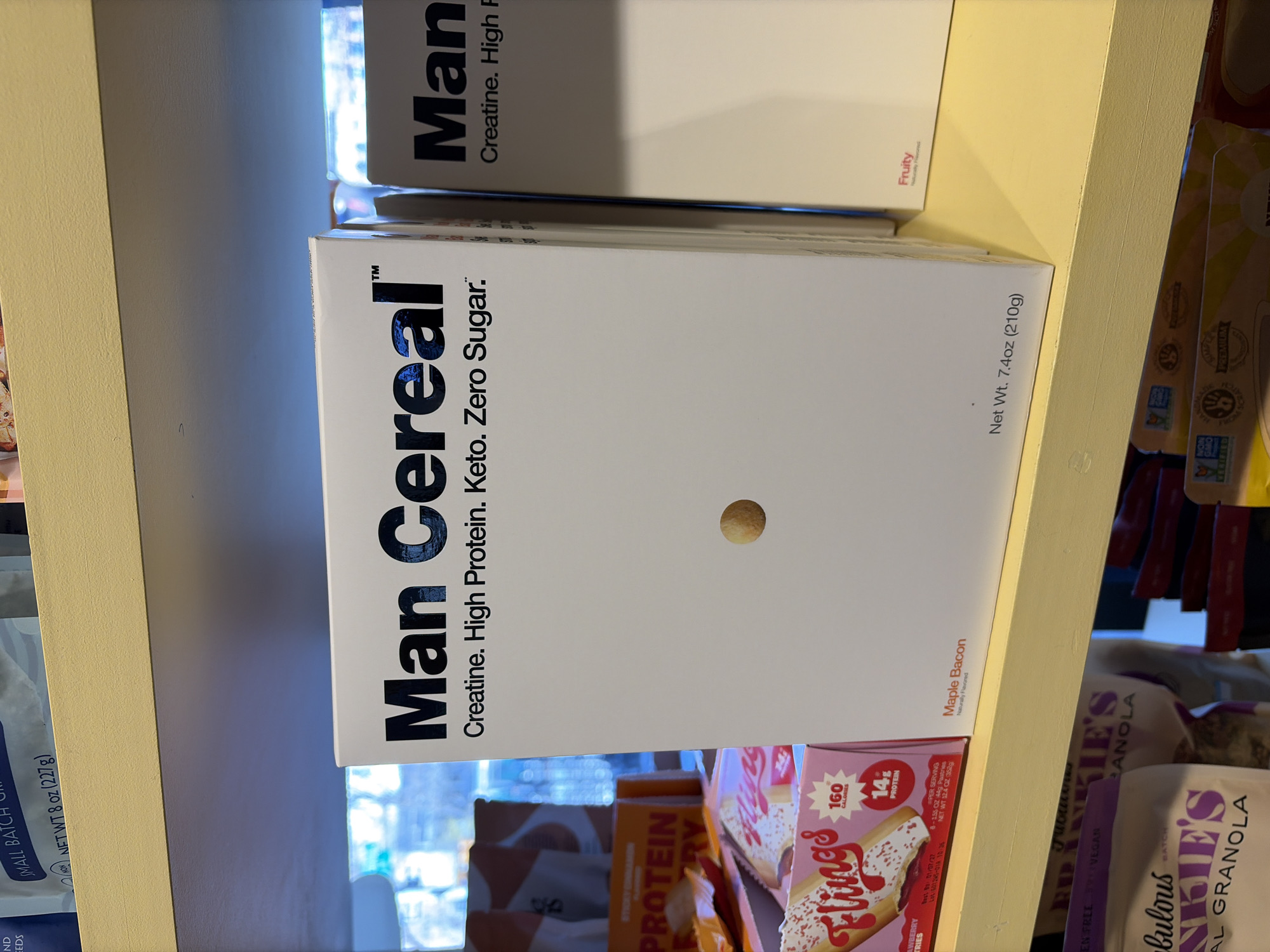 The image shows a close-up of a shelf stocked with various food products. Dominating the view is a white box of "Man Cereal" with the text "Creatine. High Protein. Keto. Zero Sugar." in black. Below this box are other food items, including boxes of "Flings" and bags of "Fabulous Kookie's" granola. The shelf appears to be made of light yellow material. In the background, there are more indiscernible food products. The setting is indoors, possibly a grocery store or pantry. There is no indication that this image is in the city of New York, United States of America.