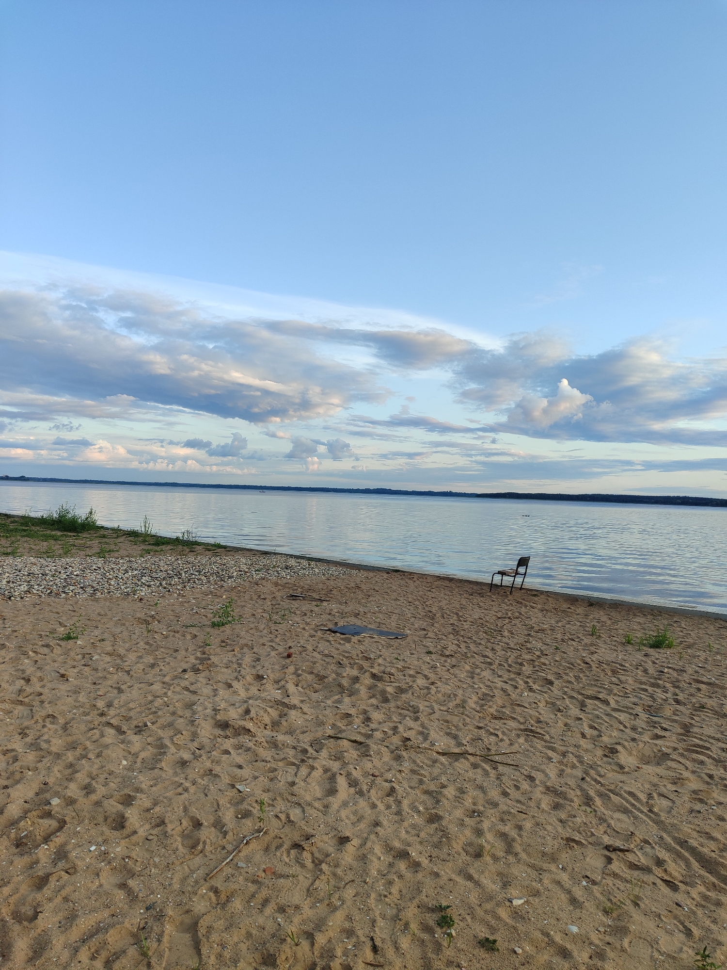 An eye-level, medium-shot photograph captures a sandy beach adjacent to a wide body of water under a cloudy, light blue sky. The foreground is dominated by a sandy beach with visible footprints and scattered pebbles. A single dark-colored chair with a metal frame is positioned on the sand near the water's edge, facing away from the camera towards the water. The middle ground features the calm water of a lake or sea, reflecting the sky. In the distance, a dark, thin line of trees or land is visible on the horizon. The sky is mostly clear with scattered, soft white clouds, particularly in the upper left portion of the frame, suggesting it might be late afternoon or early evening. There are no people visible in the image.