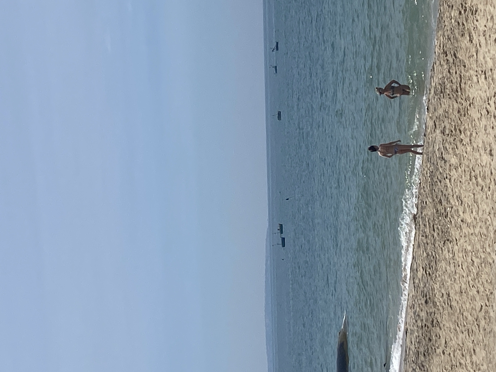A sunny day at the beach in Phu Thuy Ward, Vietnam, where two women in bikinis stand in the shallow water. The sand is a light brown color and gently slopes down to the water's edge, where small waves are breaking. The water is a calm blue, with several small fishing boats visible in the distance, as well as a landmass on the horizon. The sky is clear and a light blue, indicating fair weather.