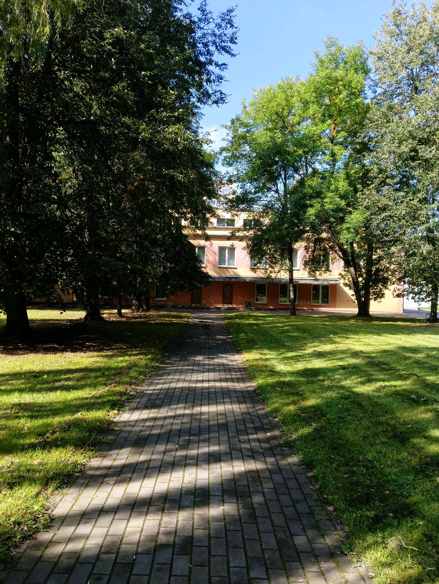 The image shows a paved walkway leading towards a light-colored, multi-story building with a pinkish-orange lower facade, set amidst a grassy lawn and mature trees. The walkway is made of dark gray rectangular pavers arranged in a basket-weave pattern, with dappled sunlight and tree shadows falling across it. The lawn is green and well-maintained, with scattered fallen leaves. The building has several windows with white frames, and its upper floors are a lighter shade, possibly yellow or cream. The trees are a mix of deciduous varieties, with lush green foliage. The sky is a clear, bright blue. The scene appears to be a park or the grounds of an institution, possibly a school or administrative building, in Hrodna, Belarus. There are no people or animals visible in the image.