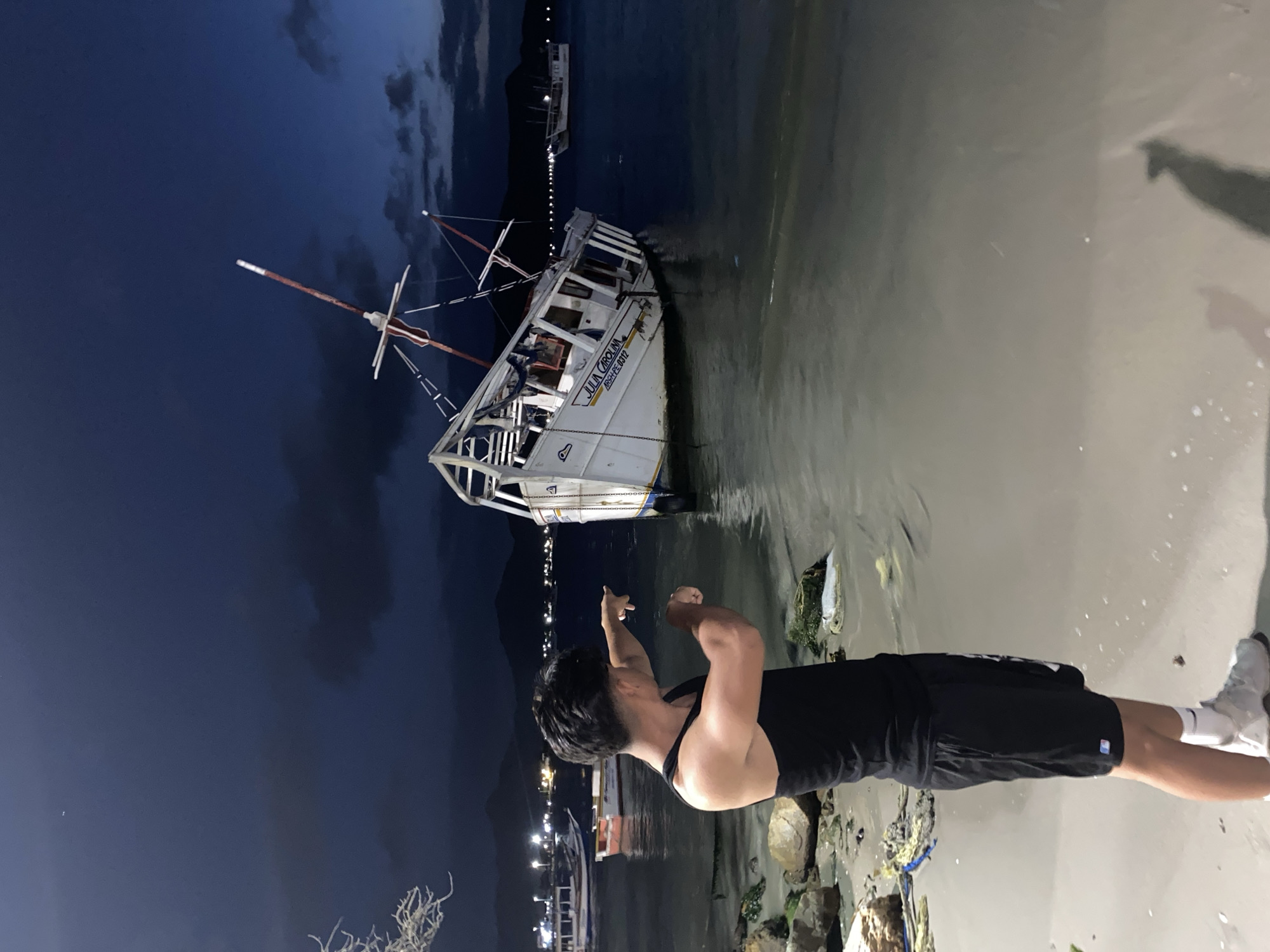 A man wearing a black tank top, black shorts, white socks, and white shoes stands on a beach. He is pointing towards a partially submerged boat. The boat is white with a red and white mast and has the name "JULIA CAROLINA" and "8559-FE-0312" written on its side. The scene appears to be at night, as indicated by the dark sky and the presence of city lights in the background. The location is Juan Griego, Venezuela, as per the prompt. The foreground consists of the sandy beach and debris. There are also lights in the distance on the left.