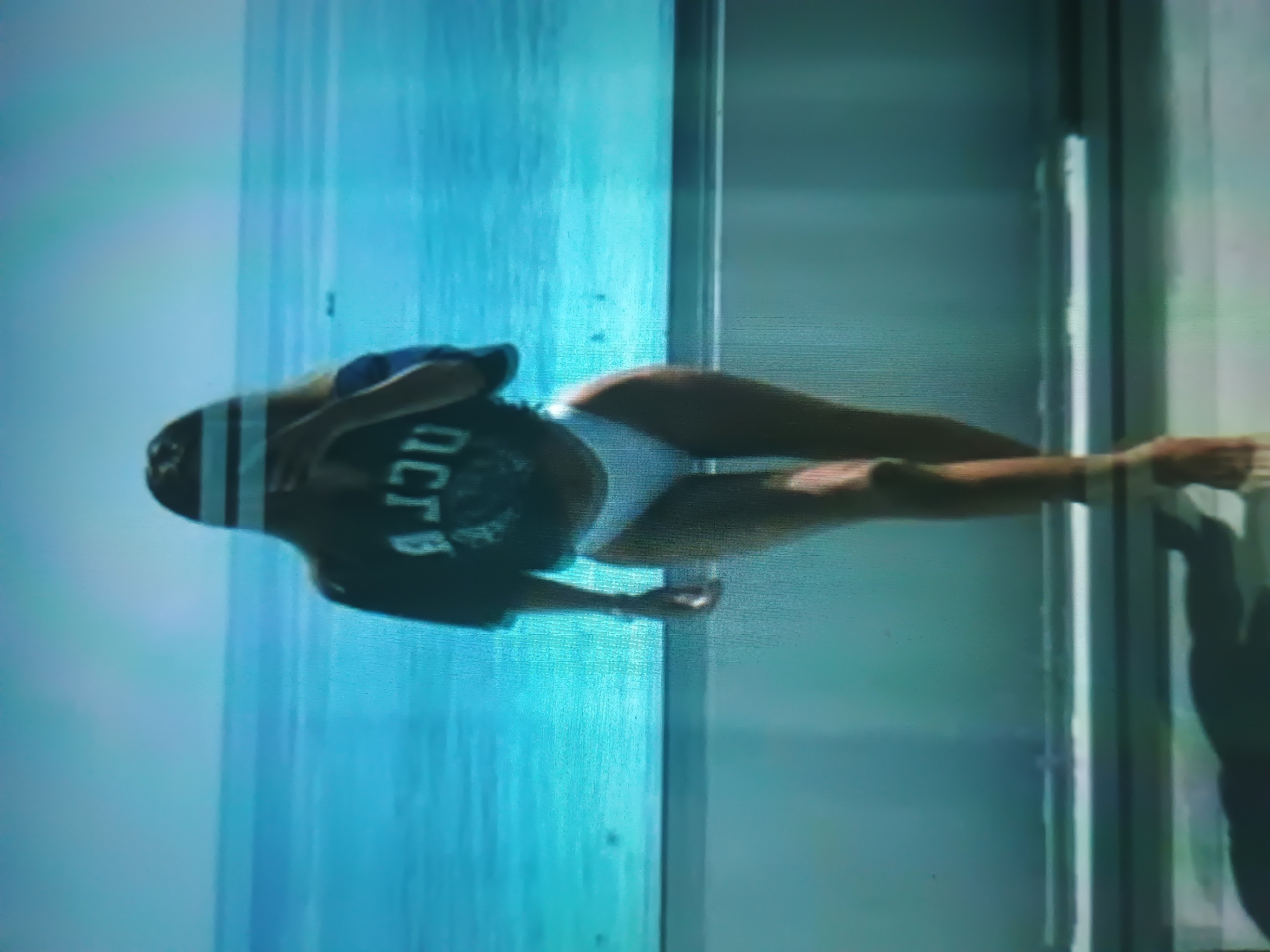 A single individual, appearing female, is depicted indoors, facing towards a large body of water visible through a window or glass barrier. The individual wears a dark, oversized t-shirt displaying the text "UCLA" in reverse across the chest, paired with white bikini bottoms. Her head and upper face are obscured by two horizontal white bars, and her right hand is partially raised towards her face. She stands with her left leg slightly forward, suggesting a walking motion. The background features an expansive blue ocean or sea under a light blue sky, with a discernible horizon line and faint dark specks on the water's surface. A horizontal rail or frame separates the indoor foreground from the outdoor view. The image quality presents noticeable scan lines and digital artifacts. The scene is an interior space overlooking a body of water.