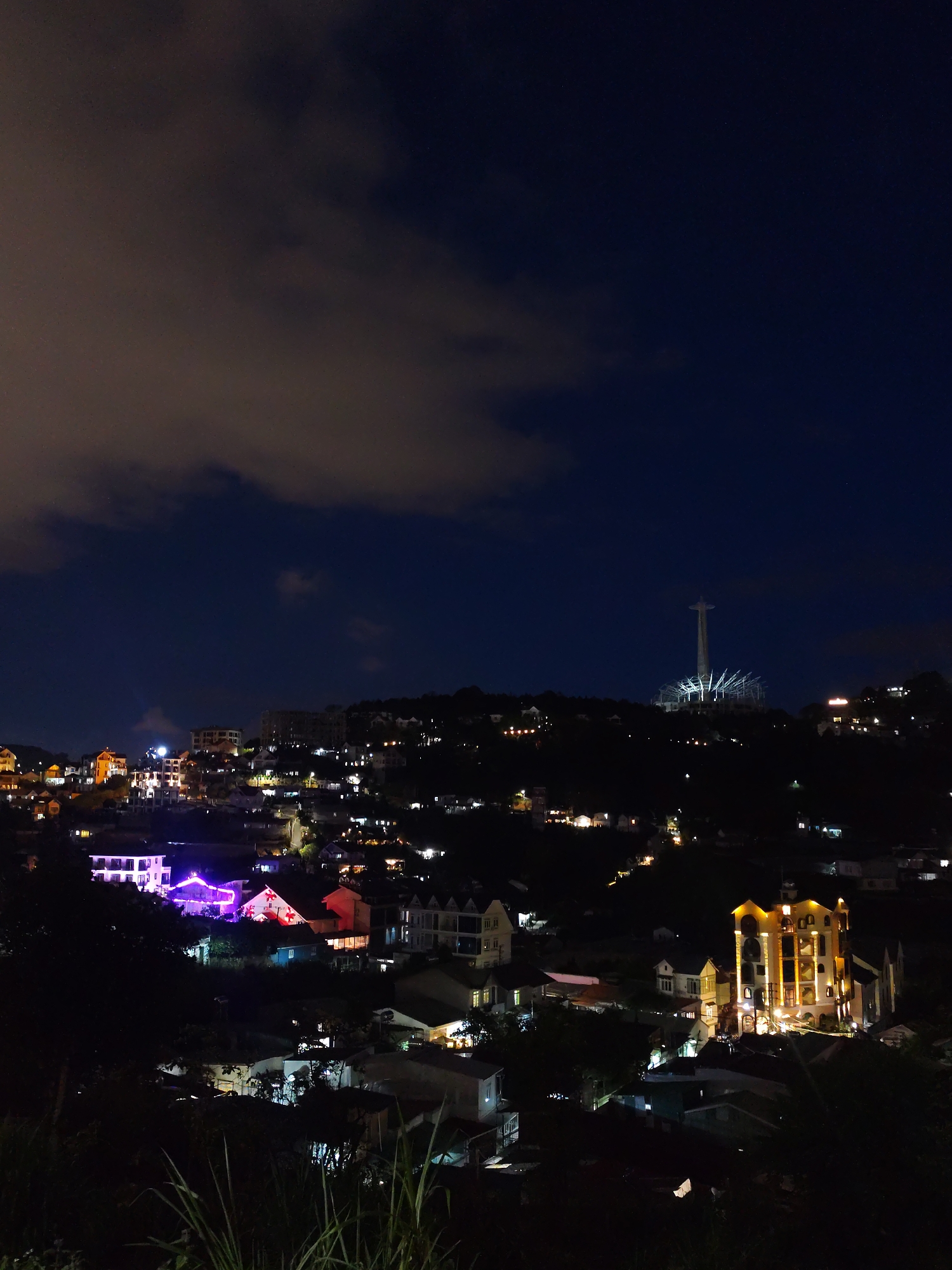 The image shows a night view of Da Lat, Vietnam. In the foreground, a residential area is visible with houses illuminated by various lights, including yellow, pink, and purple hues. A prominent building with yellow lights stands out in the right foreground. In the background, a hill with several houses and streetlights is visible. A large, illuminated tower, possibly a religious monument, is located atop the hill in the distance. The sky is dark blue with scattered clouds. The overall scene depicts a nighttime cityscape.