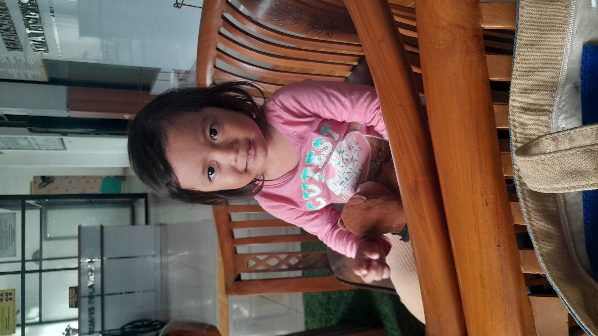 A young girl with dark hair is sitting in a wooden chair in what appears to be an indoor public space, possibly an office or waiting area in Tasikmalaya, Indonesia. She is wearing a pink long-sleeved shirt with a graphic of a cupcake and the word "CUTES" on it. She is holding a small brown teddy bear. In the background, there is a reception desk with signage that is partially visible and appears to be in Indonesian. There are also signs on the wall, one of which reads "KEJAKSAAN" and another that says "KOTA TASIKMALAYA". The overall environment seems well-lit, likely with artificial lighting, and there are some green artificial grass mats visible on the floor. A beige tote bag is visible in the foreground, partially obscuring the lower part of the scene.