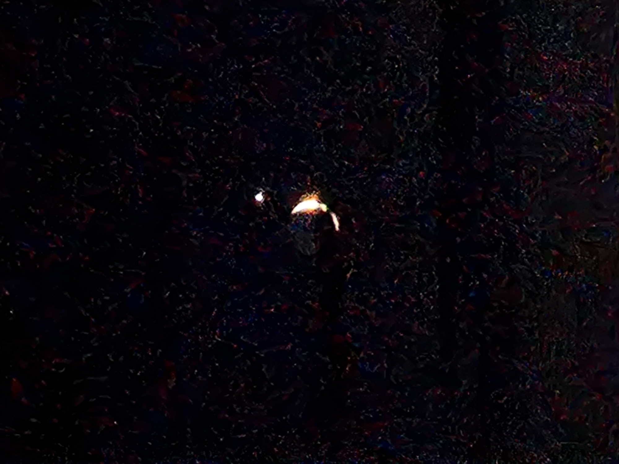 The image depicts a dark, textured background with a small, bright object near the center. The object appears to be a light source, possibly a firework or a distant light. There are hints of reddish and blue hues scattered throughout the background, suggesting a speckled or iridescent texture. The overall impression is abstract and slightly mysterious, with the central light drawing the viewer's attention in the darkness. No people, text, or other discernible objects are visible. The time of day and weather are not discernible from the image.