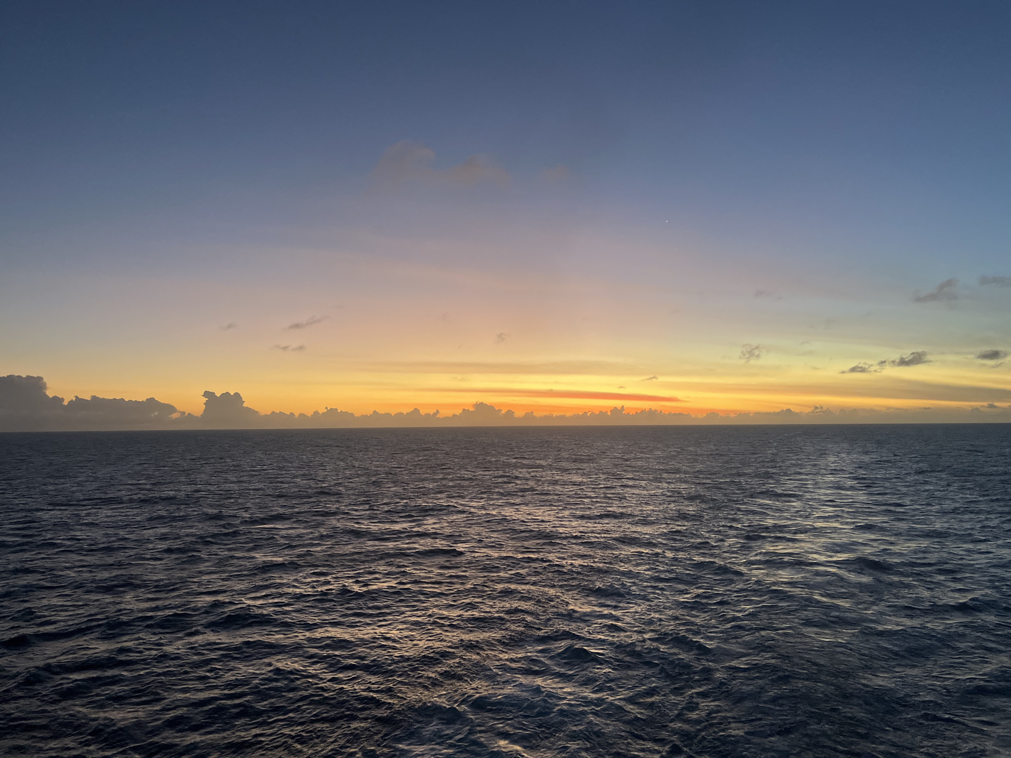 The image shows a view of the ocean at sunset. The water is dark and choppy, reflecting some of the light from the sky. The sky is a gradient of colors, transitioning from a deep blue at the top to a bright orange and yellow near the horizon. There are some small clouds along the horizon line. The location is unknown.