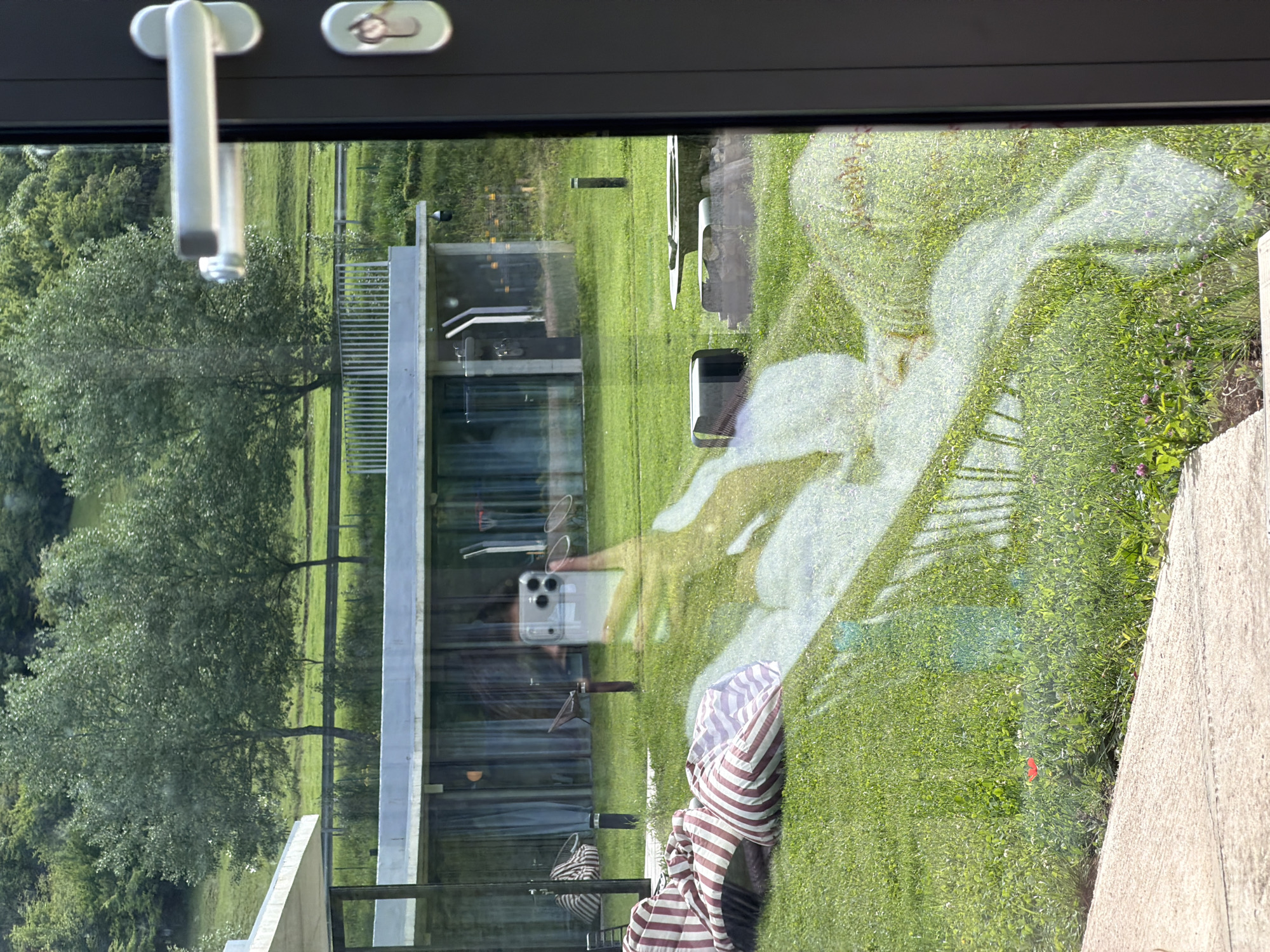 A building, a grassy lawn, and a tree are visible through a window. A modern, rectangular building with a glass facade is in the center of the frame. A manicured lawn surrounds the building, and a leafy green tree stands to the left. The reflection of a person holding a phone is visible in the glass, and a striped blanket is resting in the lower center of the frame. The window has a dark frame and a silver handle. The location is not identifiable as Chkalov, Armenia, based on the visual information.