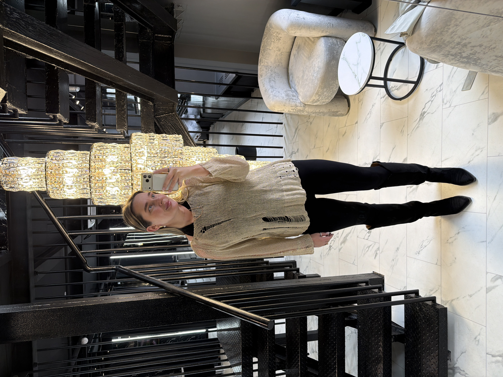 A woman is posing for a selfie on a set of black metal stairs with diamond plate treads. Above the stairs is a large chandelier with many lights. She is wearing black pants, black boots and a light-colored sweater with holes in it. Behind her is a white and grey patterned floor with two plush chairs and a small table. The location is Moscow, Russia.