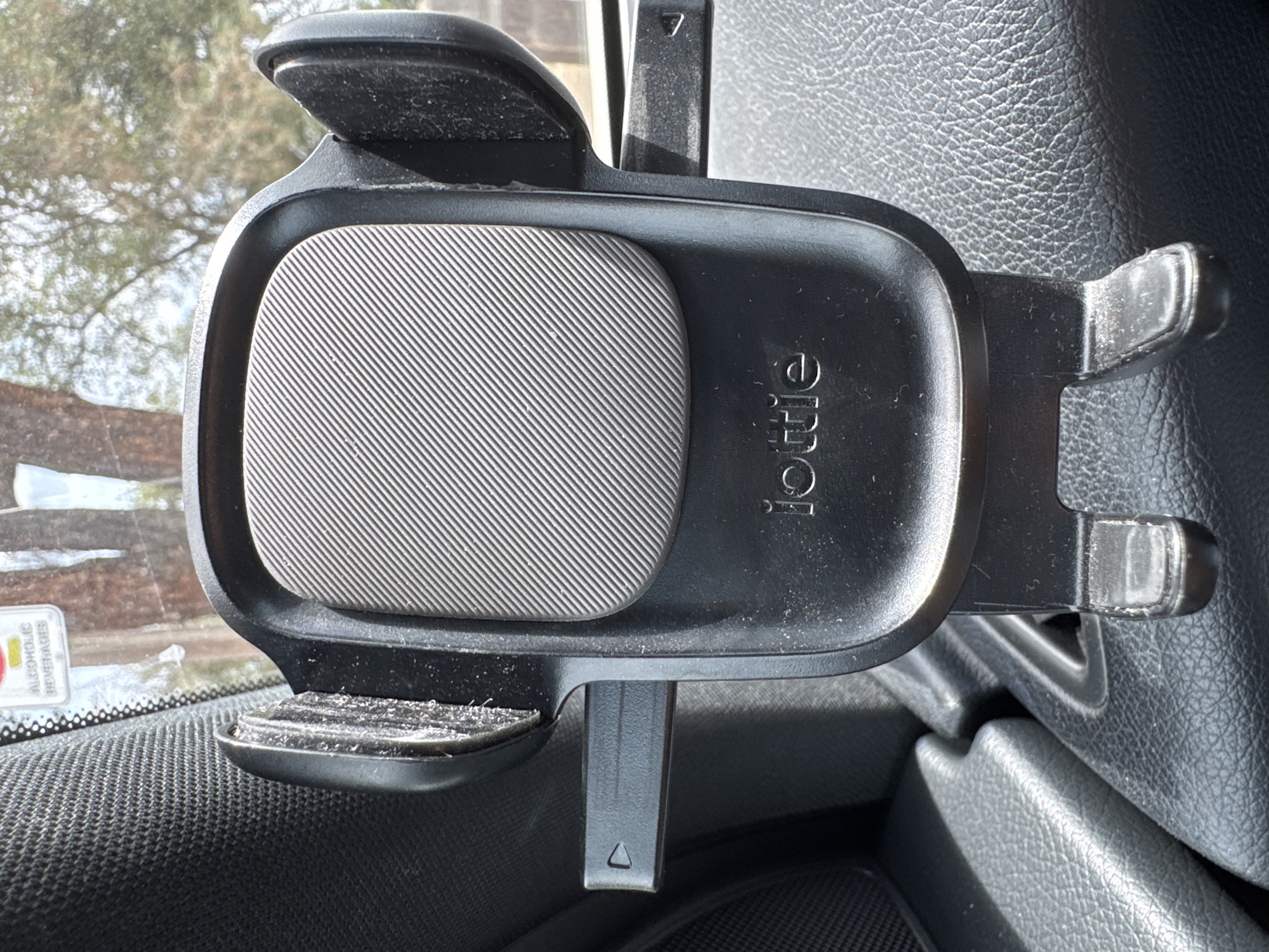 A car phone mount is mounted on the dashboard of a vehicle. The phone mount is black and has a textured gray pad for holding a phone. The brand name "iOttie" is visible on the mount. The mount is positioned in front of a windshield, and through the windshield, trees and foliage are visible, suggesting an outdoor environment. The interior of the car, including the dashboard and a textured leather surface, is also visible. There is a sticker on the windshield with red and yellow text. The scene is photographed from a slightly upward angle, focusing on the phone mount.