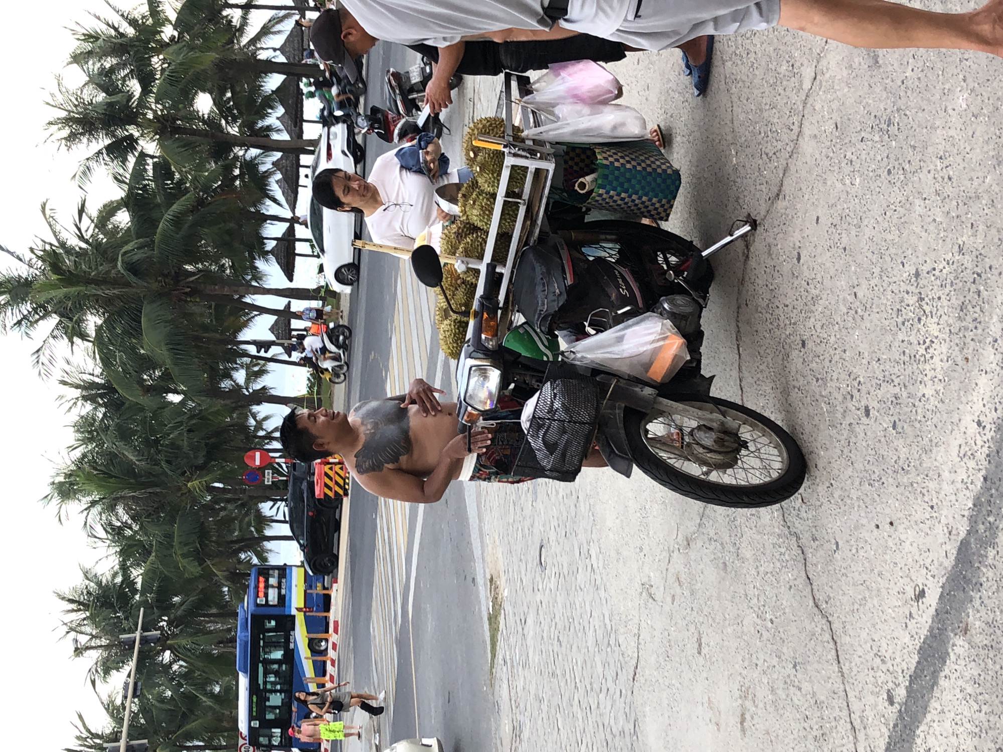 This image depicts a street scene in Đà Nẵng, Vietnam, likely near a coastal area given the presence of palm trees and the overall tropical atmosphere. A man with extensive chest tattoos, wearing patterned shorts, is standing next to a motorcycle loaded with durians. He is looking towards the right of the frame and has his hand on his chest. A woman in a white shirt and dark shorts is standing behind the motorcycle, holding a bag and a piece of fruit, possibly interacting with the man. Another man, wearing a dark cap and grey shorts, is also present on the right side of the frame, looking at his phone. The motorcycle serves as a mobile fruit stand, with durians displayed on a rack and plastic bags hanging from it. In the background, a blue and yellow bus is visible on the road, along with other vehicles and pedestrians. The ground is paved with asphalt and cobblestones, and traffic signals and street signs are present. The overall scene suggests a bustling street environment where vendors sell goods from their motorcycles.