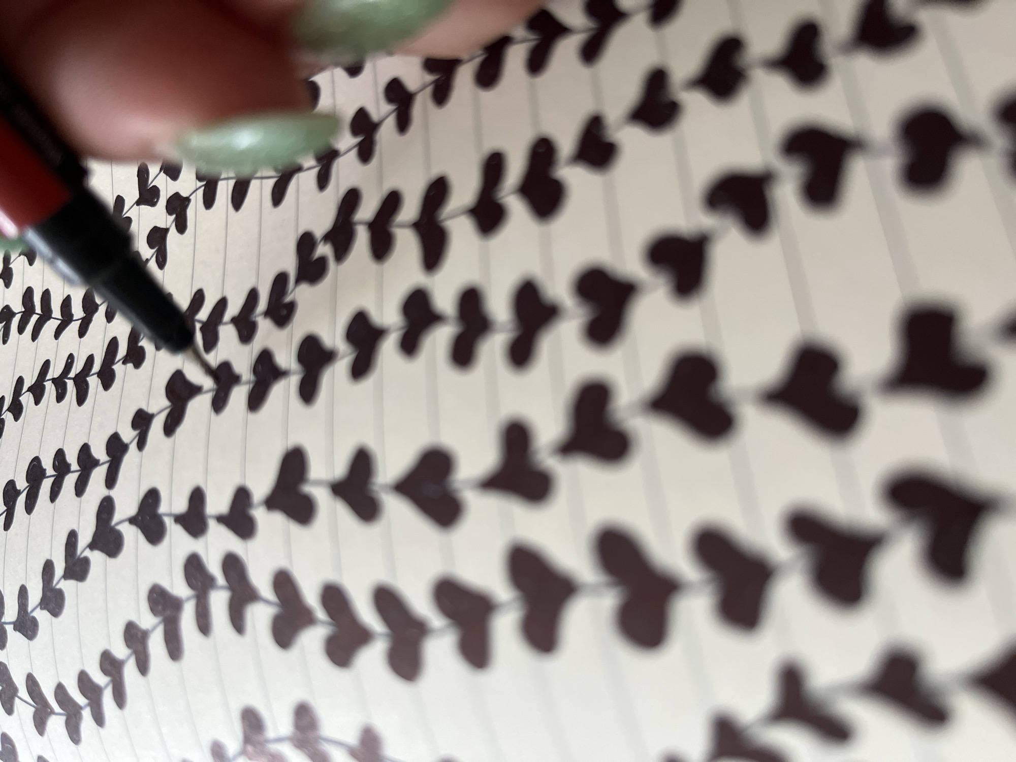 The image shows a close-up view of a hand with light green painted fingernails holding a black and red pen over a notebook with lined paper. The notebook has a repeating pattern of small, dark brown heart shapes connected by thin lines, creating a vine-like design. The pen's tip is positioned near the top of the frame, suggesting it is being used to draw or write. The background is slightly blurred, with the focus primarily on the hand, pen, and the notebook's pattern. There is no discernible information about the city of Krasnyye Barrikady, Russia, in the image.