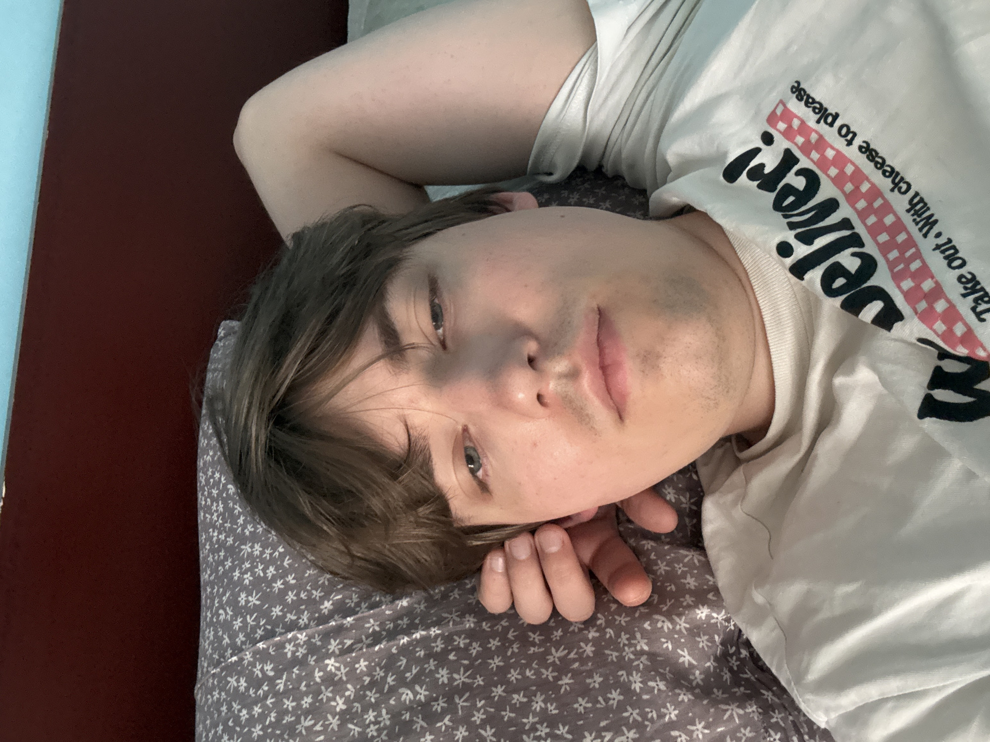 A young man with brown hair is lying on a bed, looking at the camera. He is wearing a white t-shirt with text and a checkered pattern on it. His left arm is bent and resting behind his head. The bed has a pillow with a gray floral pattern and a dark red headboard. The background shows a light blue wall. The image appears to be taken indoors, in a bedroom. There is no information available to determine the specific location in the city of Dnipro, Ukraine, or any other specific details about the environment.