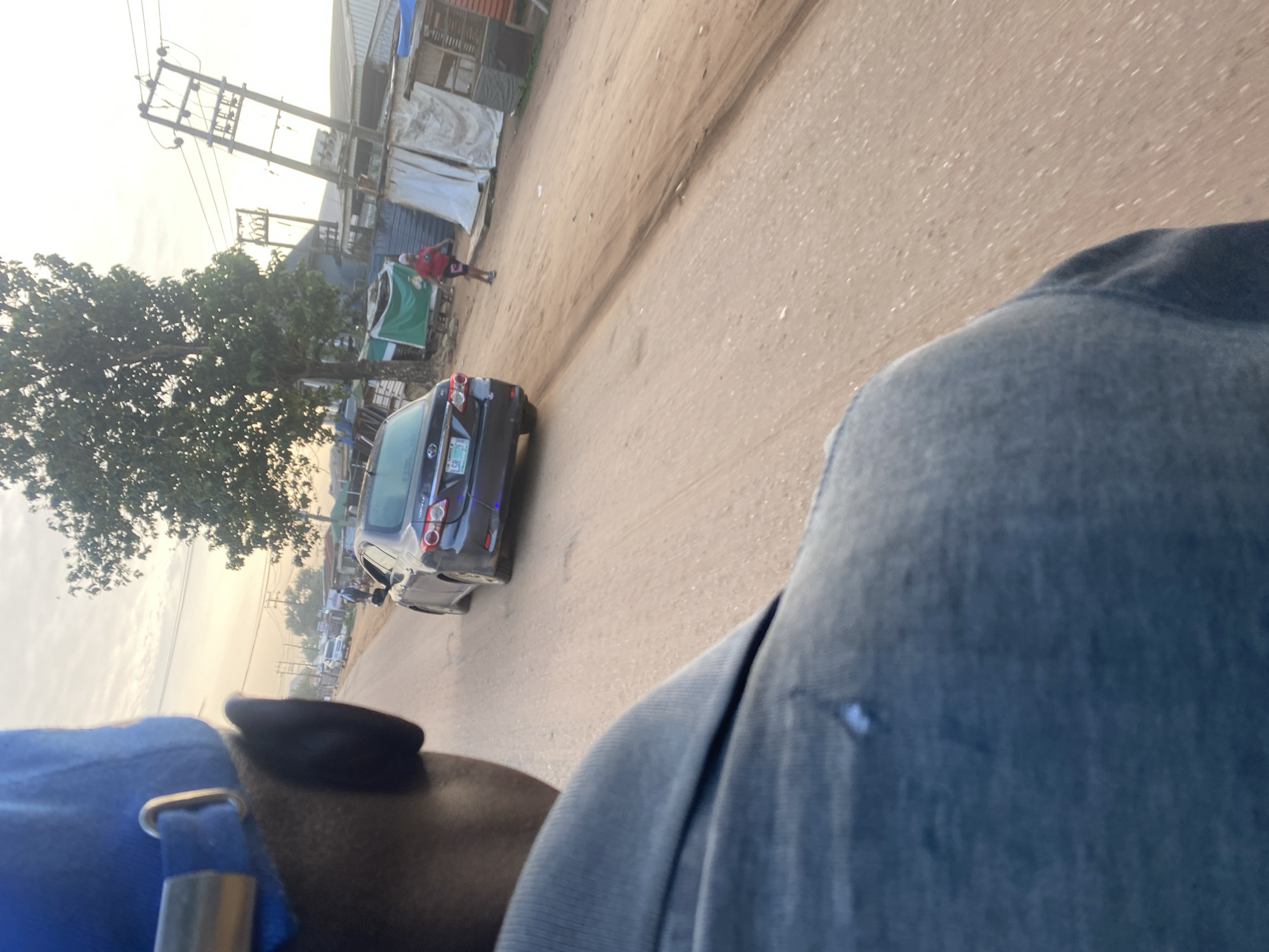 The image depicts a street scene in Ikorodu, Nigeria. In the foreground, from a first-person perspective, a person wearing a blue cap and a gray shirt is visible. The focus is on the back of their head and shoulder, suggesting they are either a passenger on a vehicle or walking ahead. In the middle ground, a dark-colored Toyota Corolla is driving on a dirt road. The car's license plate is partially visible. Further in the background, a woman in a red shirt and dark shorts is walking on the side of the road, near some stalls or informal structures. Electrical poles and wires are also present, indicating urban infrastructure. The environment appears somewhat dusty, consistent with a dirt road. The time of day seems to be late afternoon or early morning, given the soft, warm lighting and the angle of the sun, casting long shadows. The overall impression is of a bustling but somewhat underdeveloped urban area.