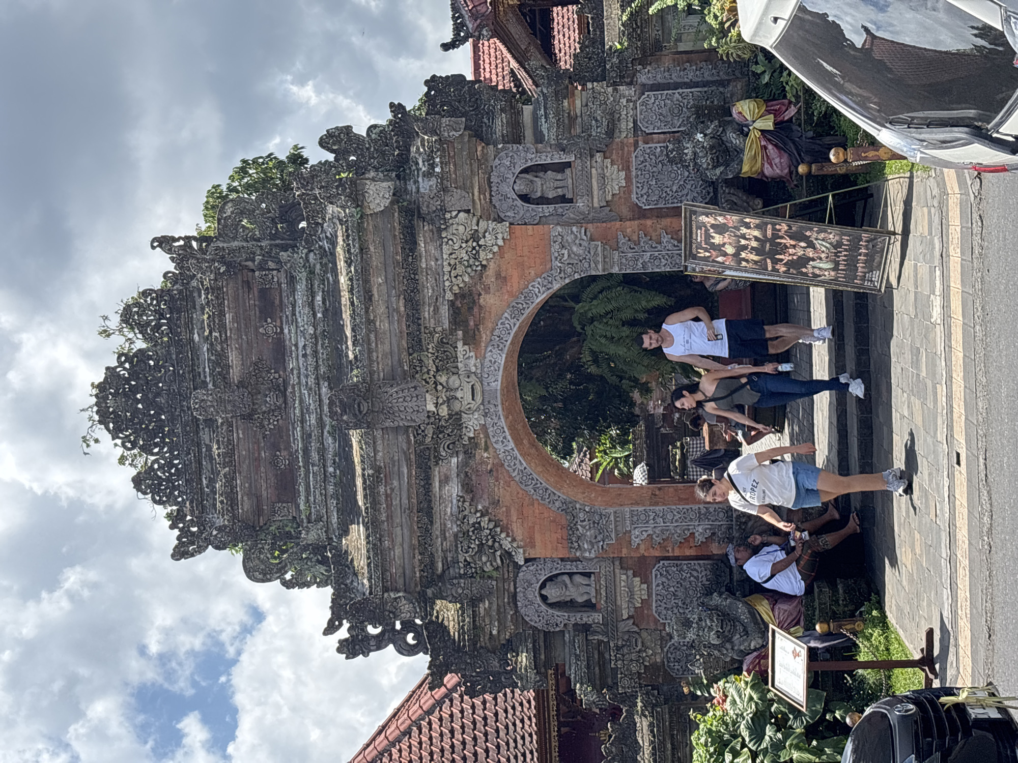 The image shows a group of people posing in front of a highly ornamented Balinese gate. The gate is made of stone and brick, and features intricate carvings and sculptures. It appears to be located in the Ubud District, Indonesia, based on the architectural style. There are four people standing and posing for a photo and one seated on the ground. Additionally, there is a sign with a frame near one of the people on the ground. A car is seen parked on the side of the road. The sky is partly cloudy.