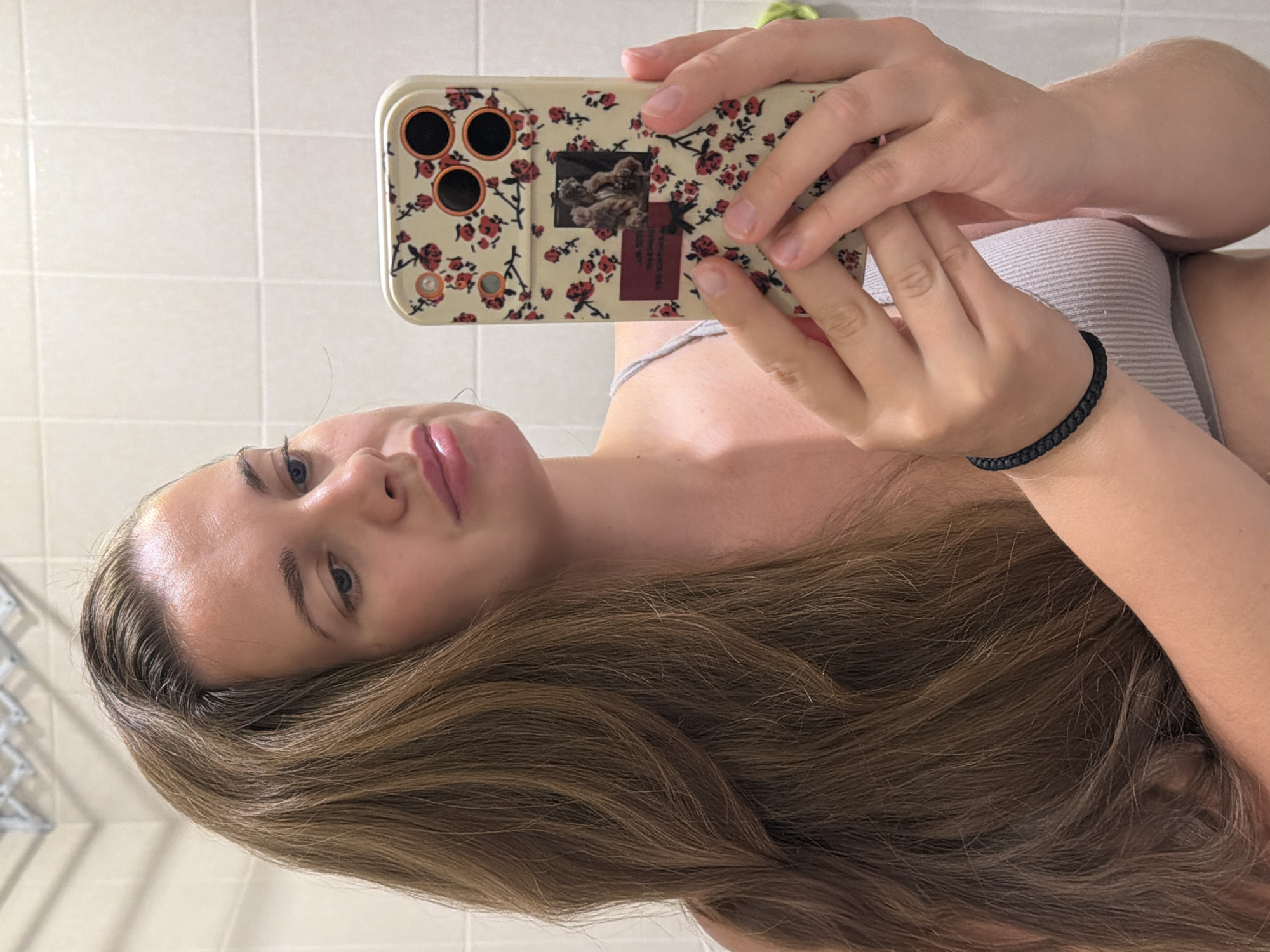 An image shows a young woman taking a mirror selfie in a bathroom. She has long, brown, wavy hair that is parted to the left and falls over her shoulder. She is wearing a light gray ribbed tank top. She is holding a smartphone in her right hand, which has a floral case with red roses and a small picture of two figures. Her left arm is bent, with her hand resting on her chest. She is wearing a black braided bracelet on her left wrist. The background is a tiled wall, likely in a bathroom. The image is captured from the chest up, with the woman's face looking towards the phone screen. The scene is a private domestic setting, typical of a bathroom in a residential building. There is no discernible information in the image that would place the location in Sochi, Russia.