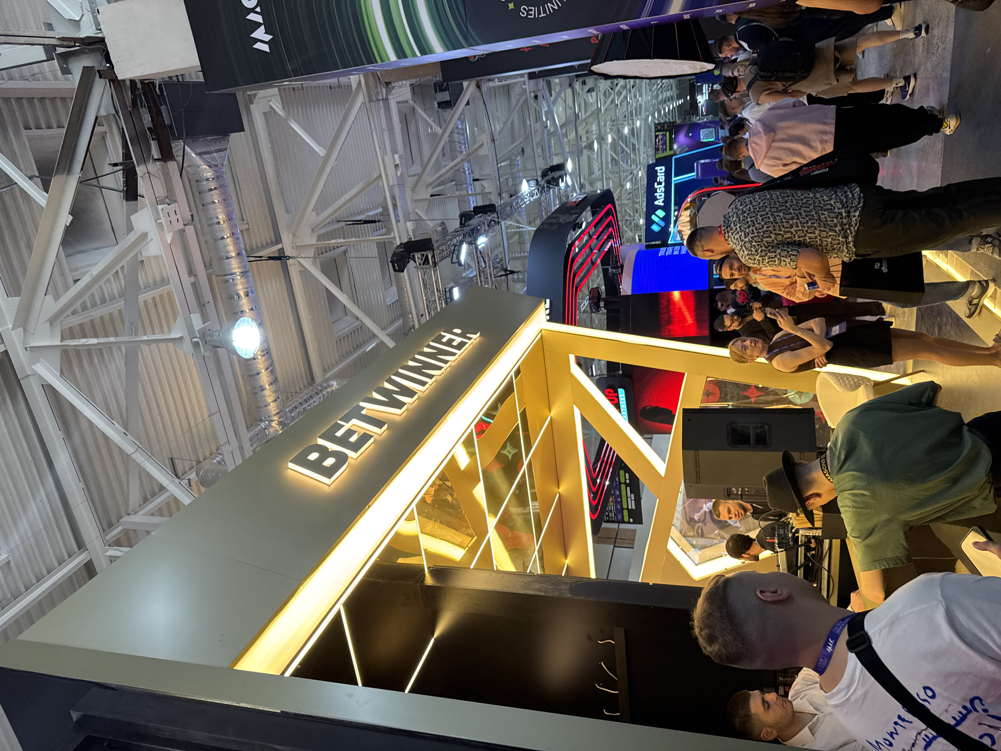 The image depicts the interior of what appears to be a trade show or convention hall, with the "BETWINNER" booth prominently displayed. Numerous people are present, engaged in conversations and interactions around the various displays. There is signage visible for "AdsCard" and other businesses. The setting appears to be inside a large building with high ceilings and complex metal beam structures. The exact location within Yerevan, Armenia, cannot be determined from the image alone.