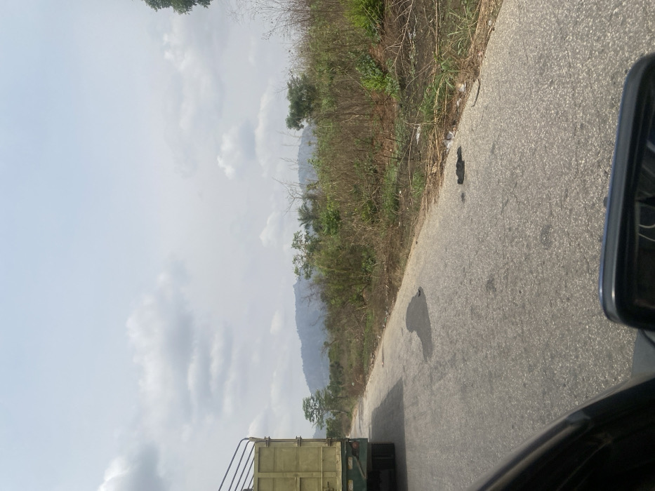 The image captures a daytime scene along a paved road in Sasaro, Nigeria, observed from the interior of a moving vehicle. In the left lane, the rear section of a light green flatbed truck, featuring high side railings, is visible. The asphalt road surface displays several dark, irregular stains. To the right of the road, a strip of sparse, dry vegetation, consisting of grasses and shrubs, extends towards a line of denser foliage and trees. A prominent range of hazy, bluish-grey mountains or hills forms the distant background beneath a sky with scattered clouds. A black rearview mirror from the observer's vehicle is partially visible in the lower right corner. No human figures or animals are discernibly present.