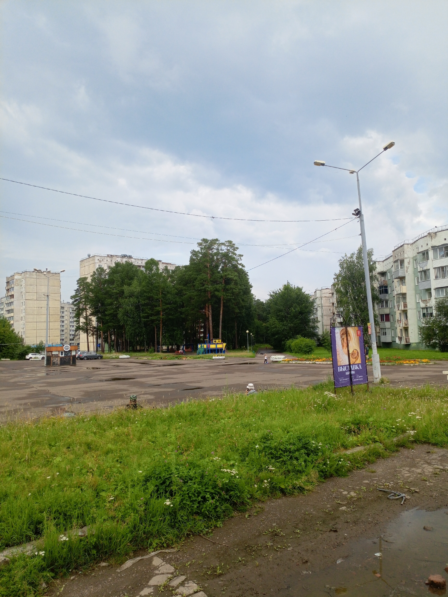 The image depicts a cloudy day scene in a residential area, likely a courtyard or parking lot, in Zheleznogorsk, Russia. In the background, several multi-story apartment buildings are visible, characteristic of Soviet-era architecture. Dense green trees, primarily pine trees, form a backdrop behind the buildings. A street lamp with two lights stands prominently on the right side of the image, near a tall pole with a security camera. A large purple banner advertising an exhibition of paintings is displayed on a stand near the street lamp. In the foreground, a patch of overgrown green grass and wildflowers borders a paved area with puddles, suggesting recent rainfall. A small, weathered kiosk is situated on the left side of the paved area, with a few cars parked nearby. A children's playground with colorful equipment is visible amongst the trees. A few individuals are sparsely present in the scene; a person is seated on the grass in the lower left, another individual is walking in the middle ground near the playground, and a third person is visible walking near the buildings in the background. Power lines are strung across the upper portion of the image, typical of urban infrastructure. The overall impression is one of a functional, somewhat utilitarian urban environment with elements of nature interspersed.