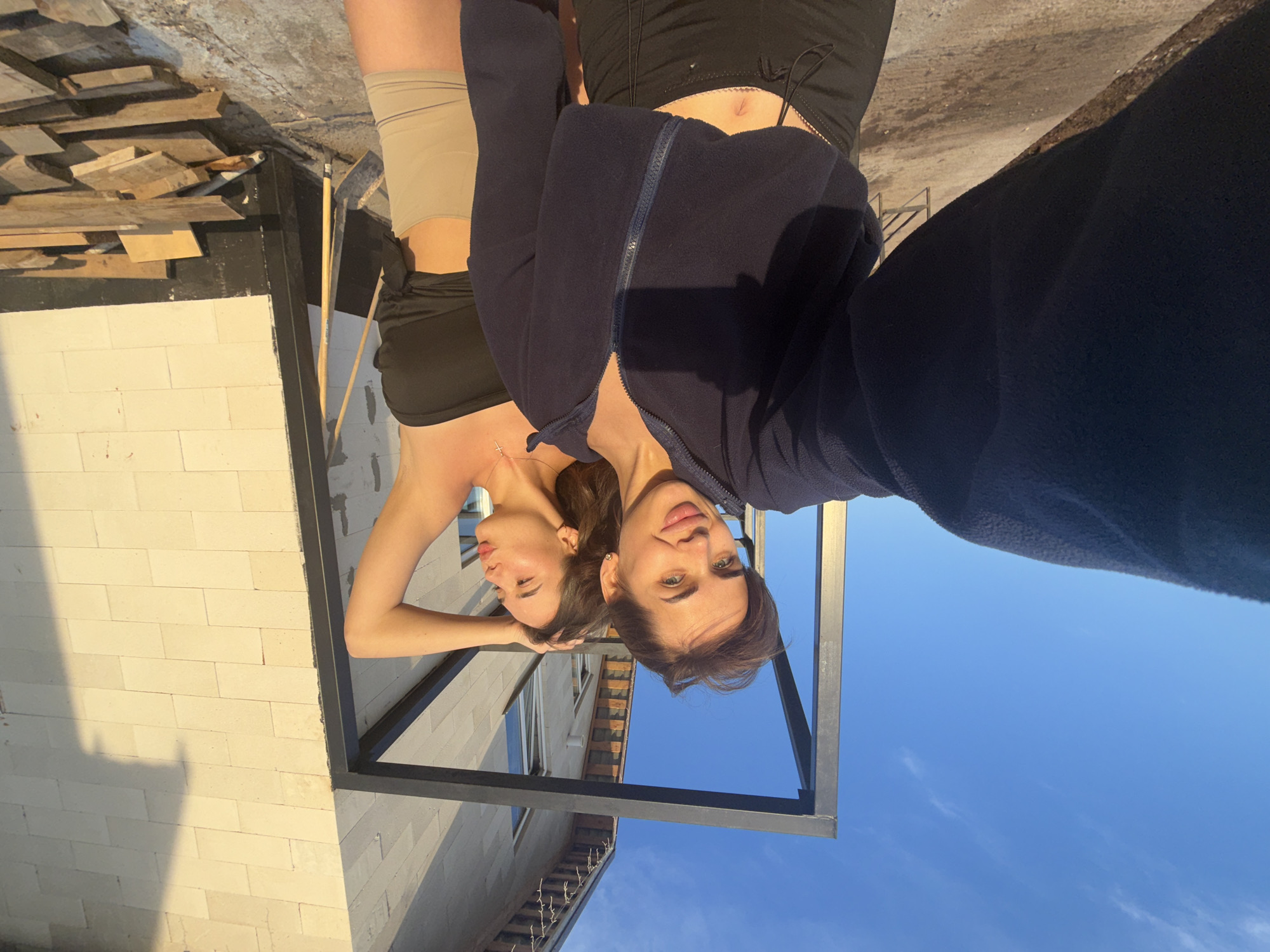 Two women stand mirrored in a black metal frame, viewed from a low angle. One woman, in a navy zip-up jacket, faces the camera directly. The other woman, wearing a black top, has her arm raised to her head. The background shows a clear blue sky and a building with white brick. The foreground includes wooden planks and construction materials. Location: Лобаново, Russia.