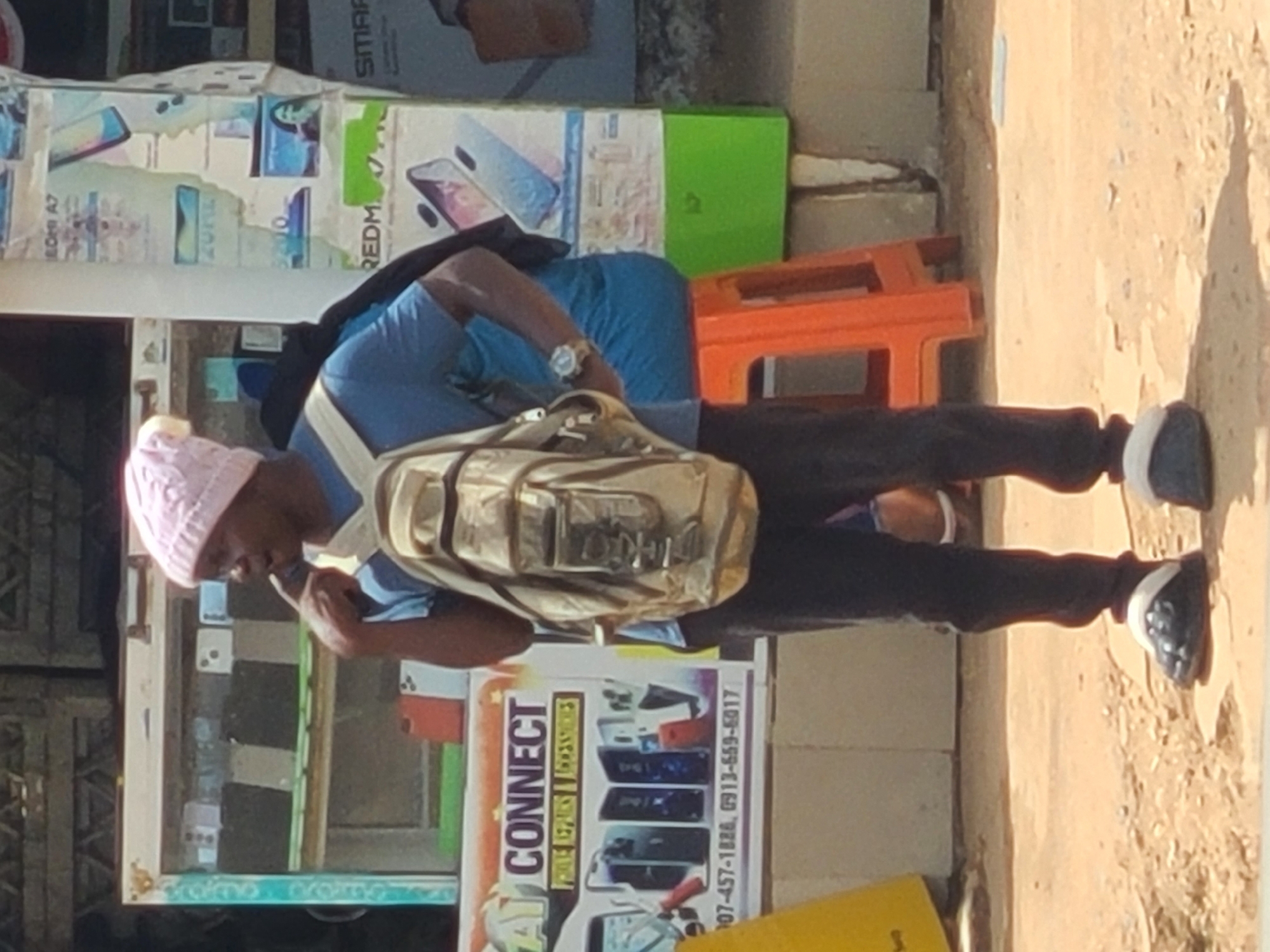 A person is standing outdoors in what appears to be a marketplace or street stall area in Jos, Nigeria. They are wearing a blue t-shirt, black pants, a pink beanie, and dark shoes with white accents. A tan messenger bag is slung across their body. The person is looking down and appears to be holding something near their mouth.
Behind the person, there is a stall with various items displayed, including what look like mobile phones and accessories. A prominent sign reads "CONNECT" in large letters, with smaller text beneath it that is partially obscured but includes phone numbers "07-457-1826, 0913-659-5017". Other posters with images of phones are visible on the stall.
To the right of the stall, an orange plastic stool is visible, suggesting a seating area. The ground is paved with concrete and dirt. The lighting suggests it is daytime, with shadows cast on the ground. The overall scene depicts a commercial environment, likely a street vendor selling electronics.