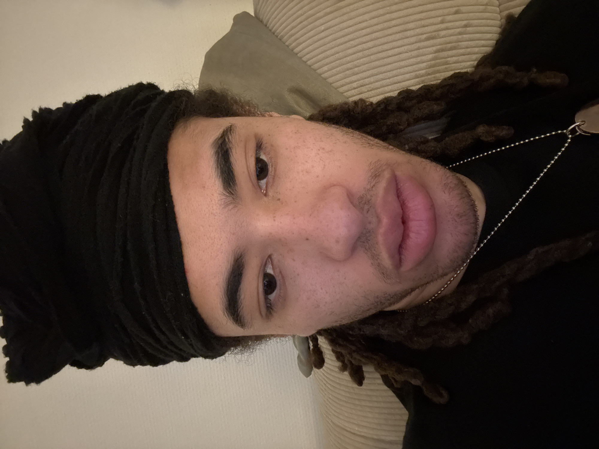 A close-up, frontal portrait features a young adult male in an indoor setting within Paris, France. The subject wears a black, textured headwrap and has dark dreadlocks visible below it. His face, illuminated by artificial light, shows medium skin tone with visible freckles/moles, dark eyebrows, dark eyes, and light stubble on the chin and cheek areas. His lips are slightly parted, and his gaze is directed downward. He wears a black long-sleeved garment and a silver-colored ball chain necklace with a small, flat, oval pendant. The background consists of a light-colored, vertically ribbed fabric object, possibly a sofa or cushion, to the subject's right, and a plain light wall with a darker, soft object (cushion) to his left. The individual is positioned centrally within the frame, occupying most of the visual space.