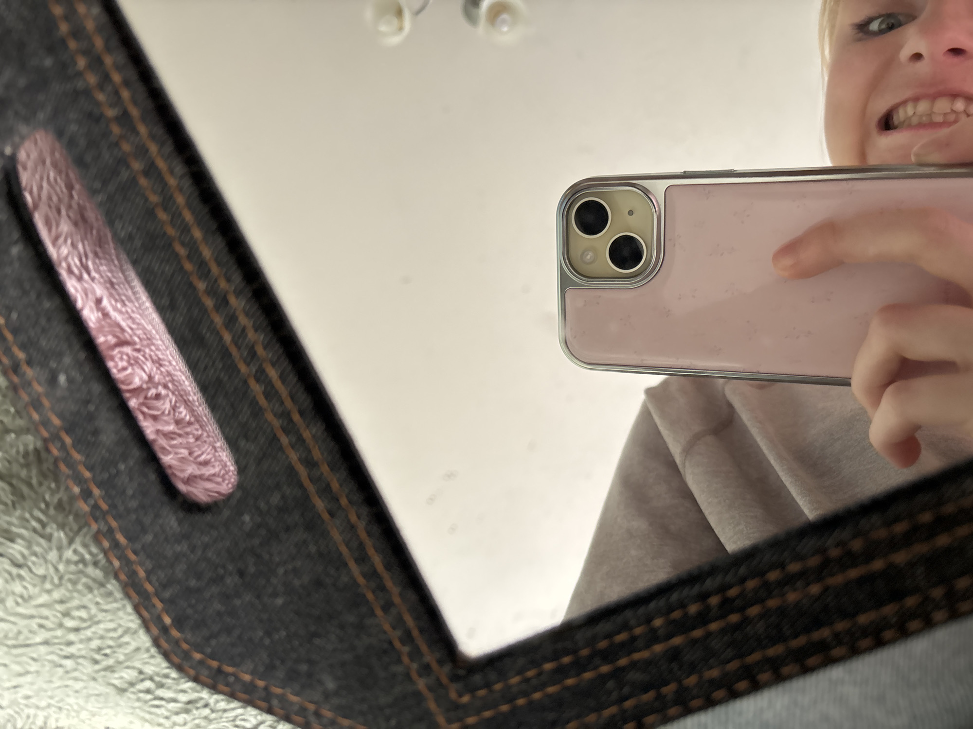 A low-angle, close-up, mirror selfie captures a person holding a pink-cased smartphone, reflecting themself in a mirror. The smartphone has a silver trim around the camera lenses, which are black. The person's face is visible, smiling with their mouth open, and blonde hair is visible. Their right hand is holding the phone, with a thumb positioned on the back. The background of the mirror shows a white surface with a few dark smudges. To the left of the mirror is a dark denim-like material with orange stitching, and a fuzzy pink object is visible in the upper part of the frame. A soft, textured grey material is also present in the upper left corner. The image is set in an indoor location, likely a bedroom, with no discernible urban context or specific landmarks to indicate a location within "Unknown, Russia".