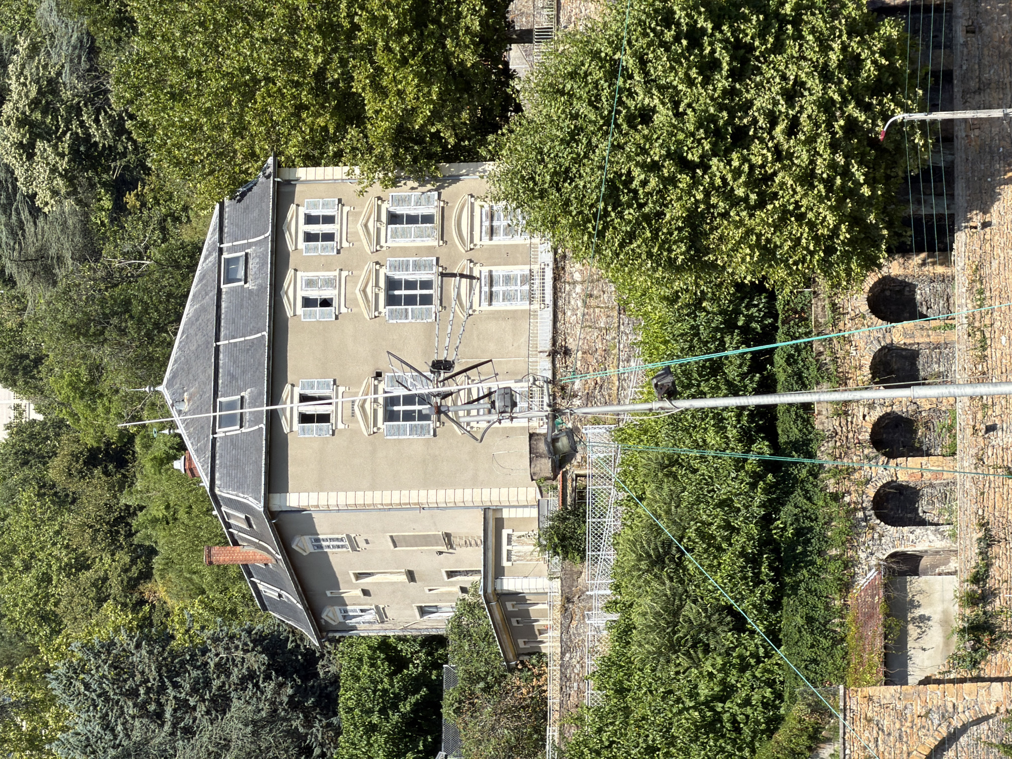 The image shows a tall, multi-story building with a grey facade and a dark, mansard roof. The building is situated on a hillside, surrounded by lush green trees. The facade features numerous windows with white shutters, and decorative architectural elements. A chimney with red bricks is visible on the roof. In the foreground, a stone wall with arched openings and greenery is present. A modern metal pole with electrical wires extends upwards through the scene, partially obscuring the building. The scene is bathed in bright sunlight, suggesting a clear day. The architecture of the building, particularly the mansard roof and the classical window surrounds, is characteristic of 19th-century Parisian or Lyonnaise architecture. Given the terraced hillside setting with stone retaining walls and the specific architectural style, this building is likely located in the Croix-Rousse district of Lyon, France, known for its steep slopes and historic buildings, particularly those associated with the city's silk industry. There are no people or animals visible in the image.