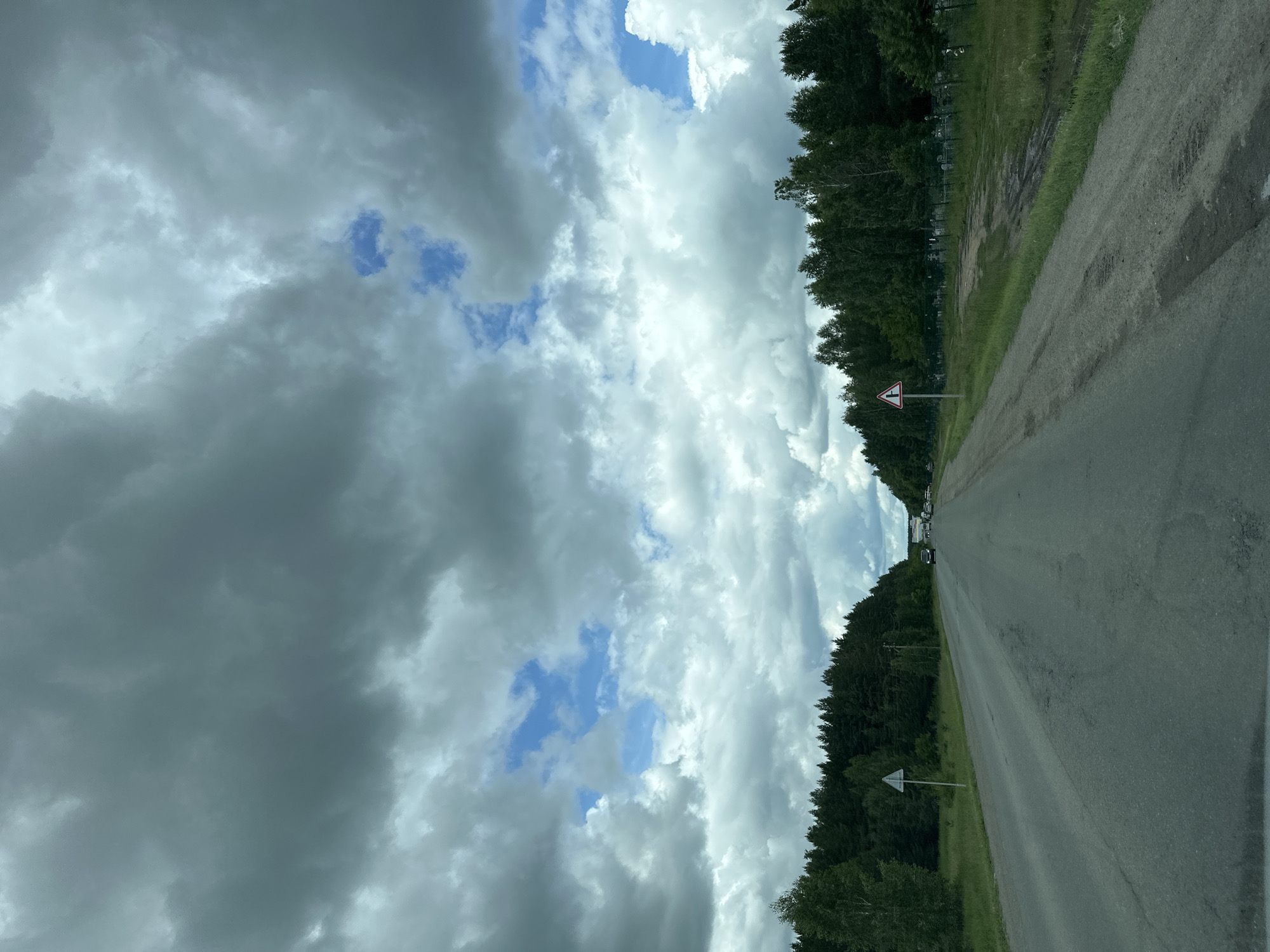 The image depicts a two-lane asphalt road stretching into the distance, bordered by dense coniferous and deciduous forests on either side. The sky above is filled with dramatic, dark storm clouds, interspersed with patches of bright blue sky. Several cars are visible on the road ahead, suggesting traffic. Road signs, including a triangular warning sign with a red border and a white interior indicating a hazard, are present along the roadside. The terrain appears somewhat uneven, with grassy verges and some areas of exposed earth. The overall impression is a view from a moving vehicle on a highway through a wooded area under a turbulent sky. The location is identified as Uvel'skiy, Russia.