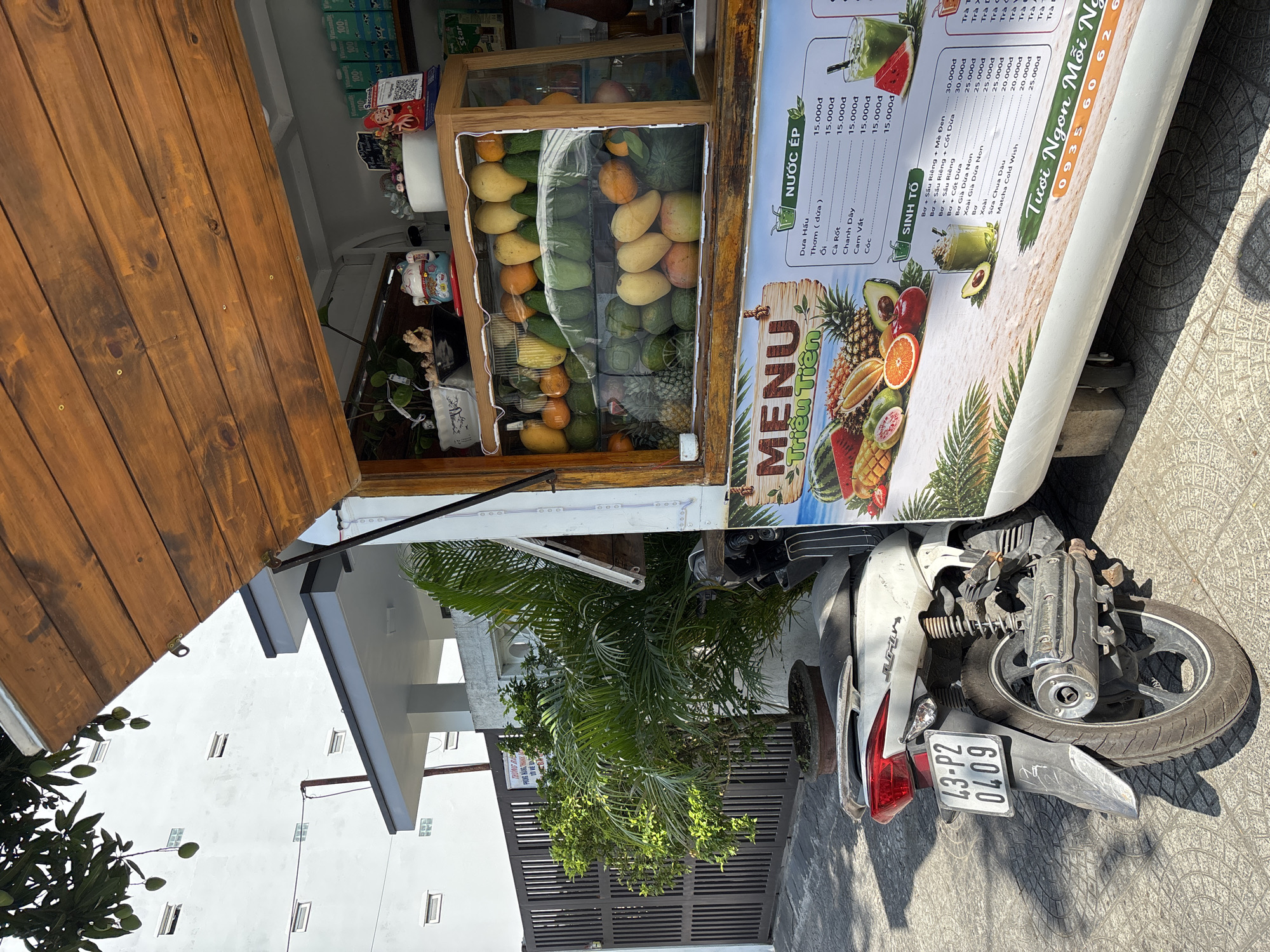 The image captures a fruit juice stall in what appears to be a residential area of Đà Nẵng, Vietnam. A motorcycle is parked in front of the stall, which features a wooden structure, a display case filled with various fresh fruits, and a menu sign with prices. The menu features the name "Triều Tiên," along with "Tươi Ngon Mỗi Ngày." The display case contains mangoes, oranges, pineapples, and other fruits. The motorcycle has the license plate "43-P2 0409." The buildings behind the stall have white walls and windows. A plant in a pot is to the left of the motorcycle. The stall’s menu displays a variety of fruit juice options. The environment is sunny, casting shadows on the ground.