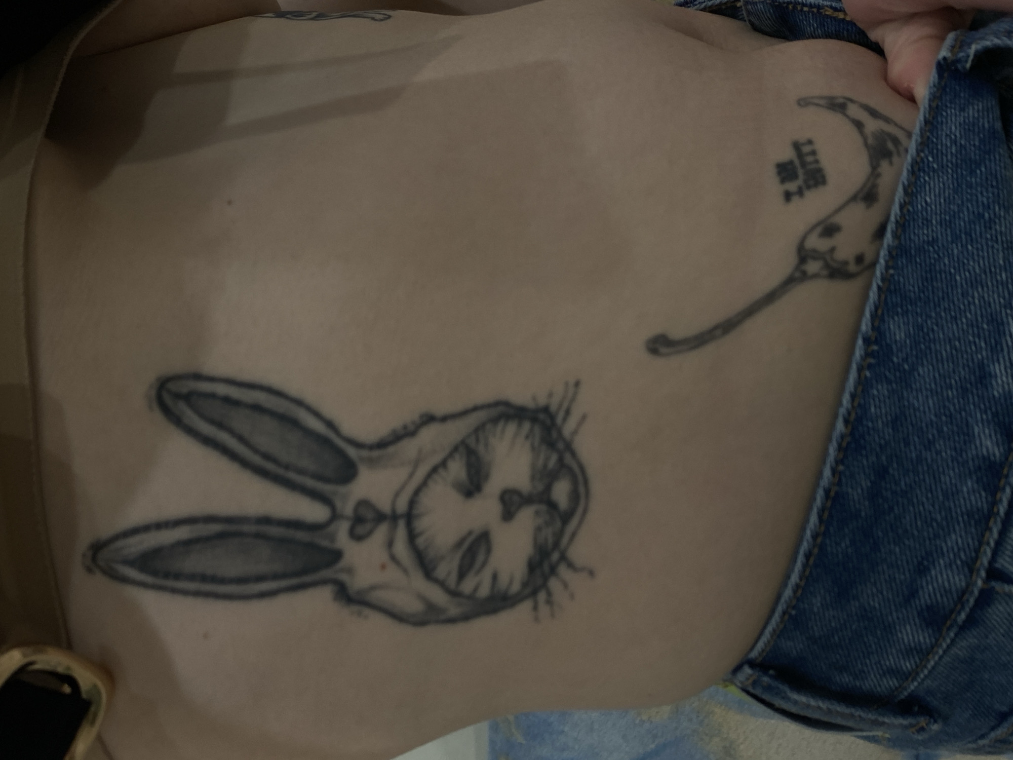 A person's torso is shown, displaying two distinct tattoos. One tattoo features a stylized image of a rabbit face enclosed within a rabbit-ear outline. The second tattoo depicts a chili pepper with text next to it. The person is wearing denim jeans with a visible gold-colored buckle or accessory near their left side. The setting appears to be indoors. There is no evidence to indicate the location being in the city of Kazan, Russia.